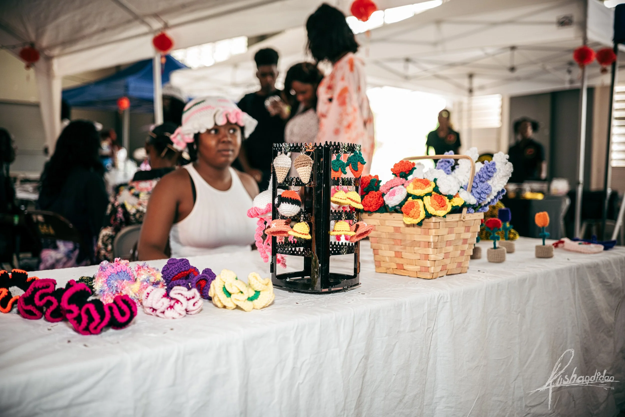 A woman sitting at a table displaying various handmade crocheted jewelry and accessories with a basket of colorful flowers, under a white tent at an outdoor market.