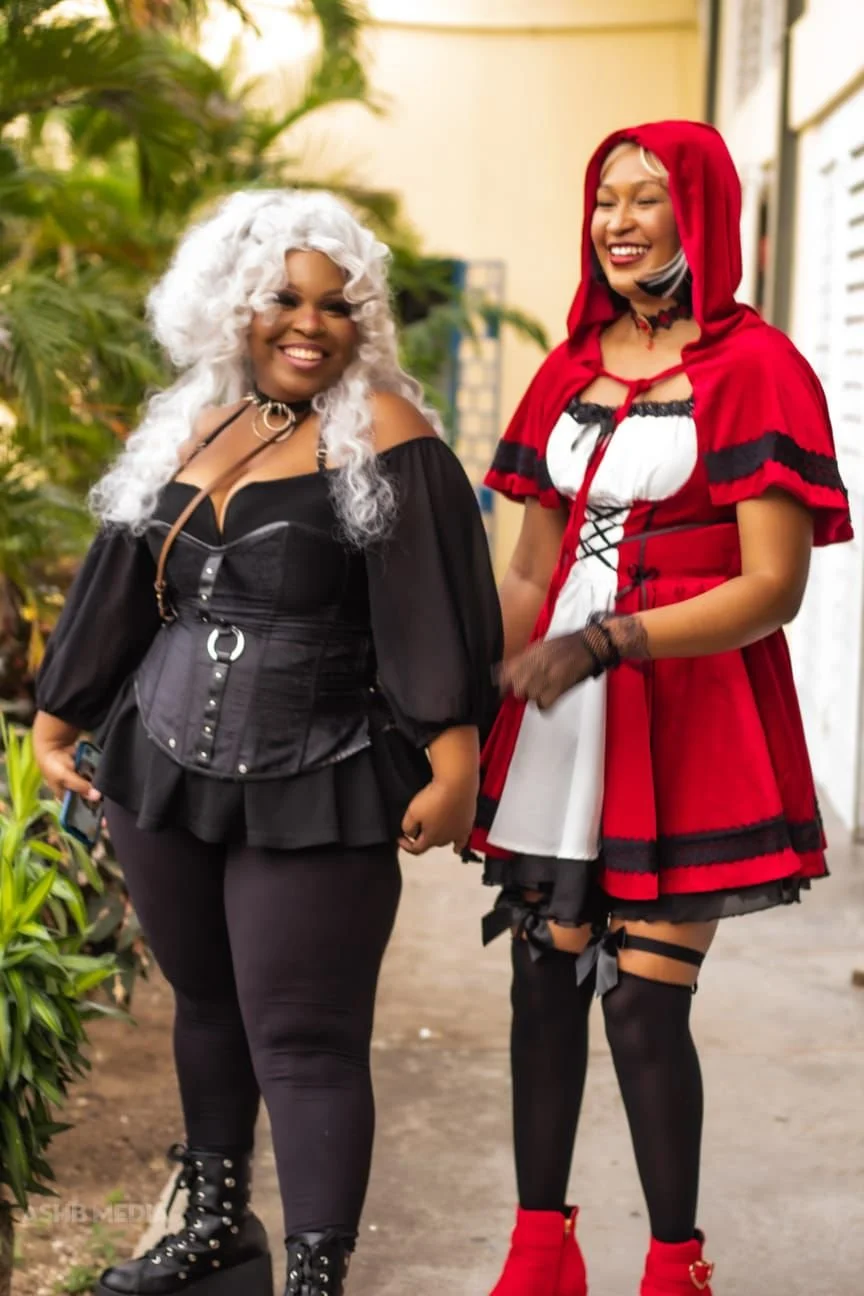 Two women dressed in Halloween costumes, standing outdoors with greenery in the background. The woman on the left has white curly hair, a black gothic dress, and black boots, while the woman on the right has blonde hair, a red hooded cape with black 