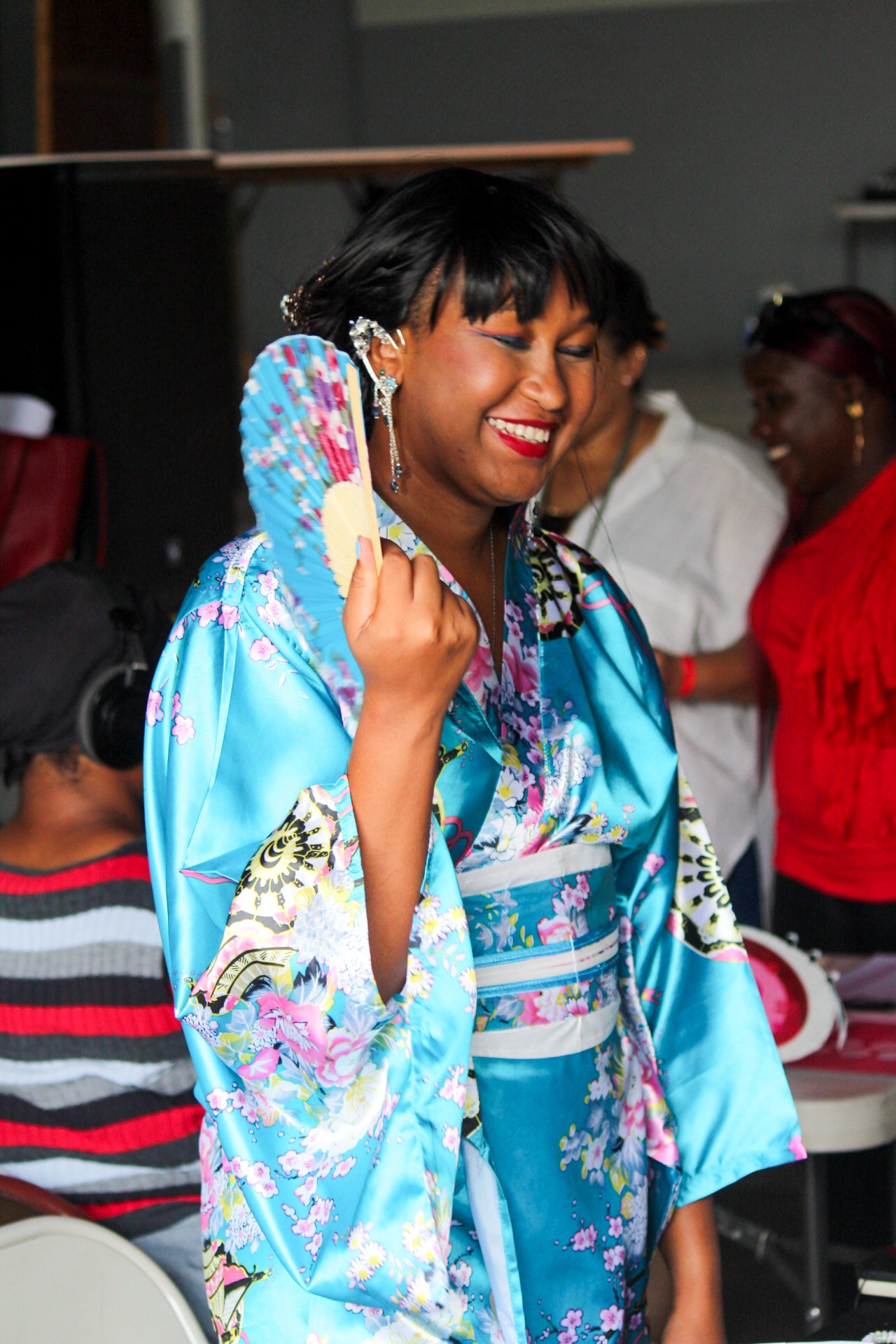 A woman in a blue floral kimono is smiling and holding a colorful folding fan. Behind her are other women, one in a striped shirt with headphones and colleagues dressed casually, in a lively indoor setting.