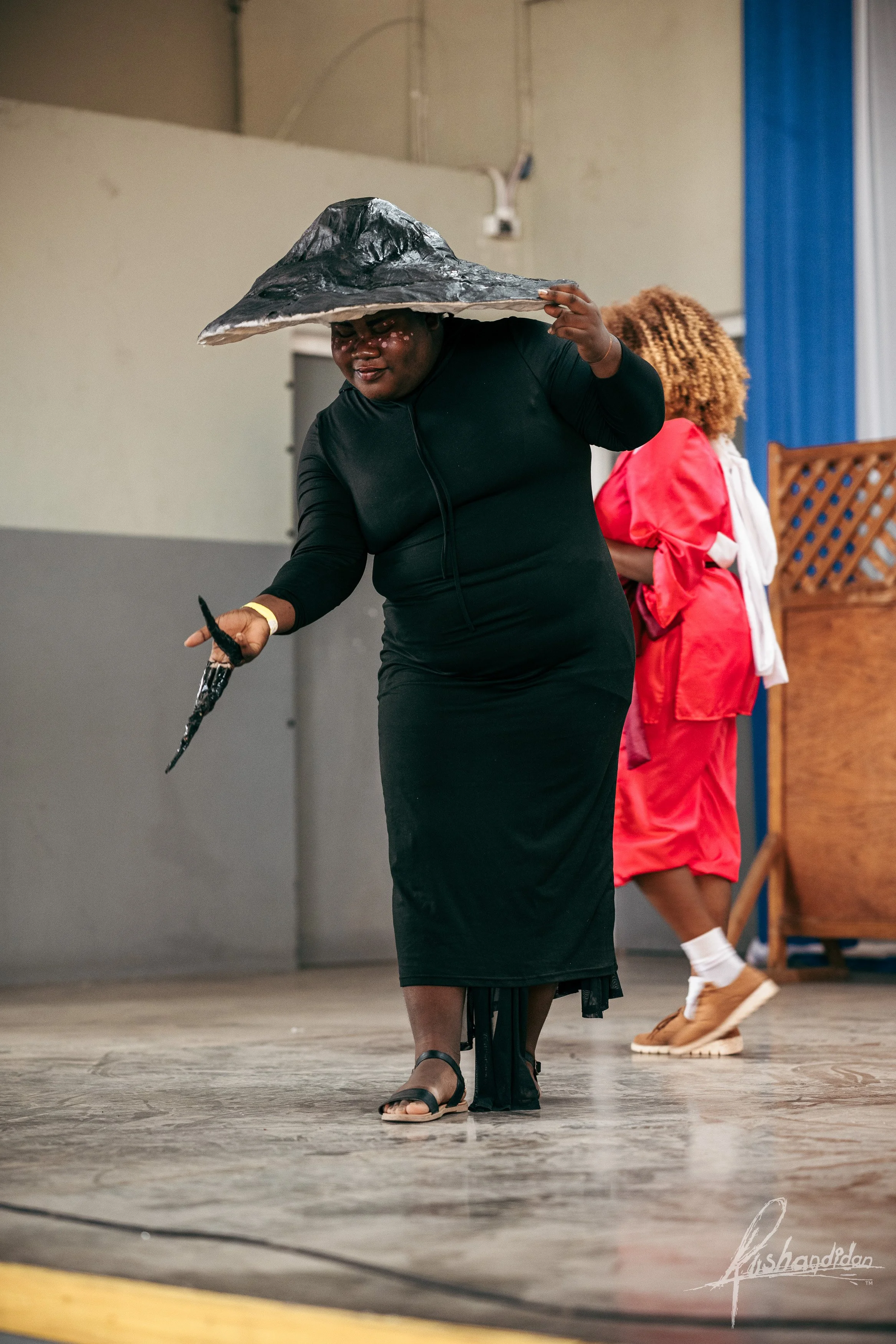 Person wearing a witch's hat, black dress, and sandals holding a wand, walking on stage with another person in a red outfit in the background.
