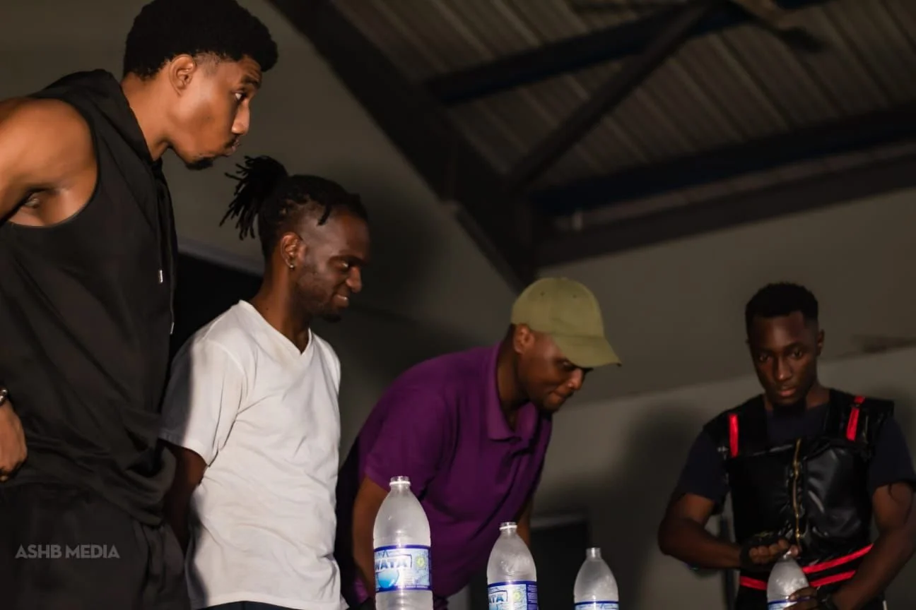 Four young men standing around a table with bottled water, indoors with a wooden ceiling. They are looking down at something on the table, appearing focused or serious.