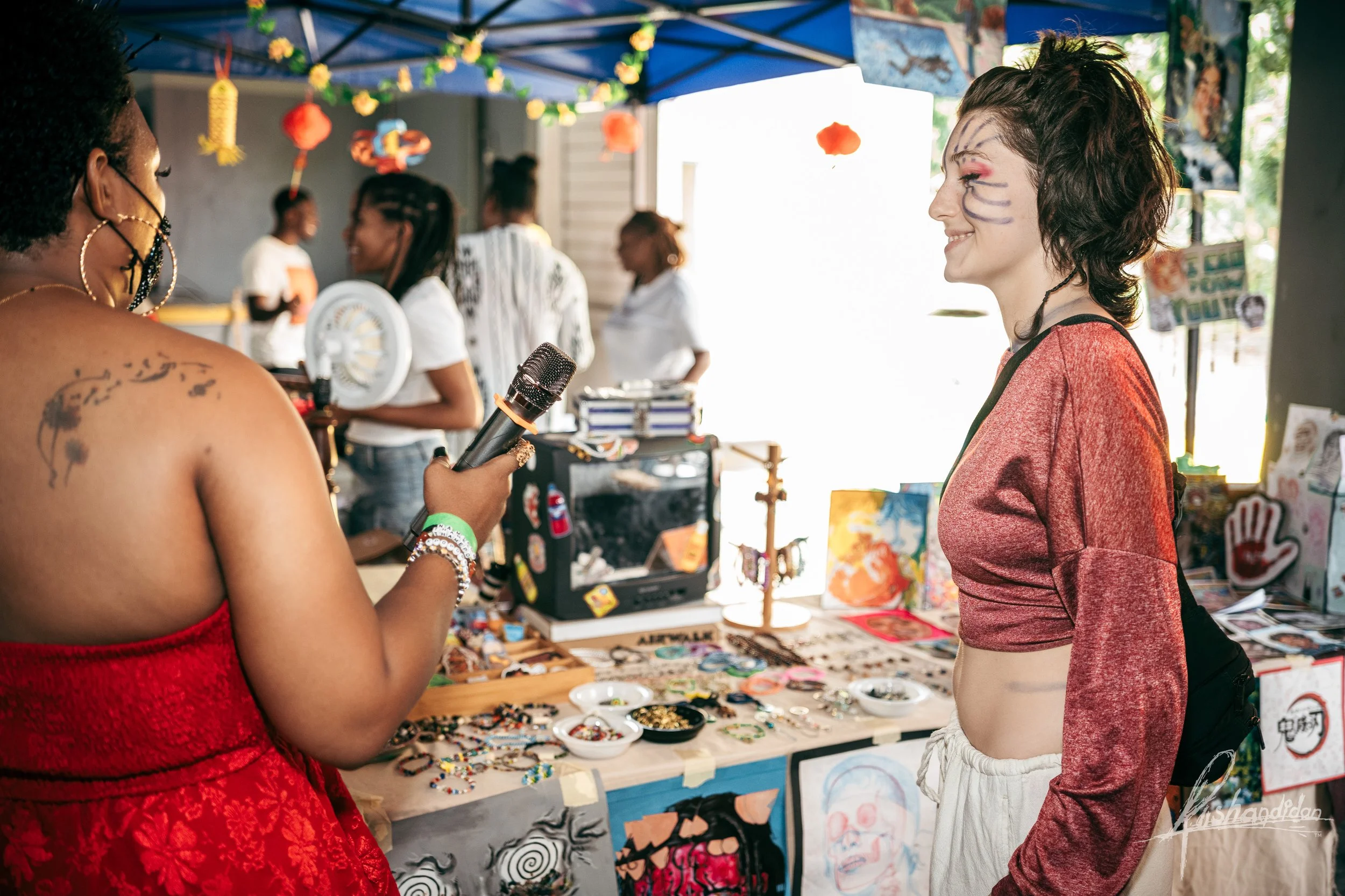 A woman with face paint and a red long-sleeve shirt shopping at a market stall, while another woman, wearing a red strapless dress, holds a microphone and talks to her. The market stall has jewelry and artwork display, with other people in the backgr