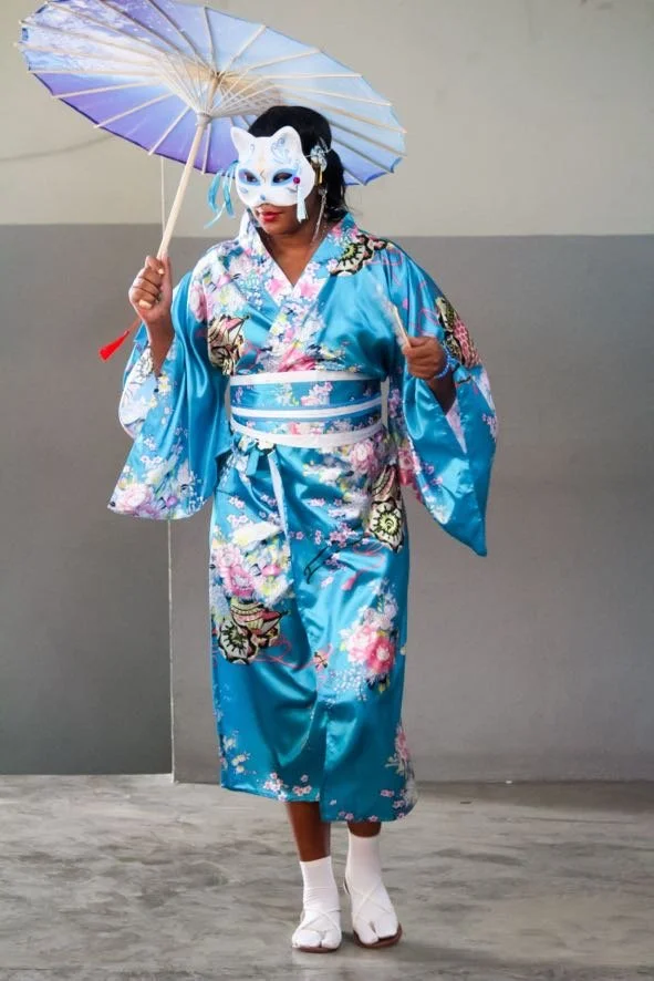 Person dressed in a blue kimono holding a traditional parasol and wearing a fox mask, standing on a plain floor against a two-tone wall.