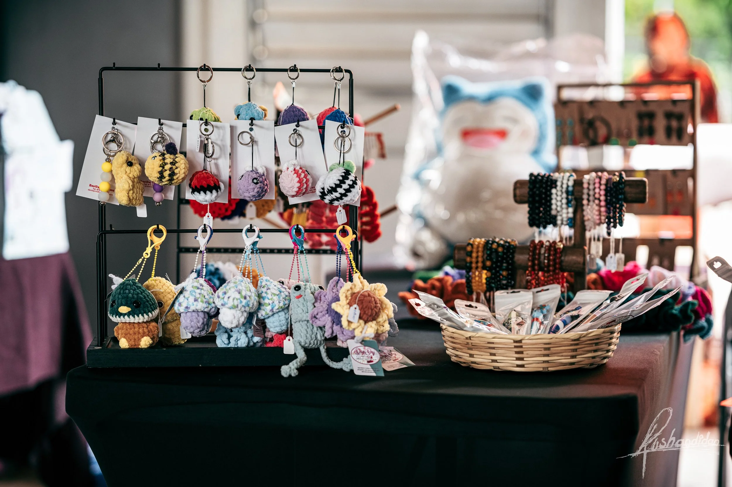 Display of handmade crocheted keychains, jewelry, and accessories at a craft fair booth, with a basket of packaged items and a plush toy in the background.