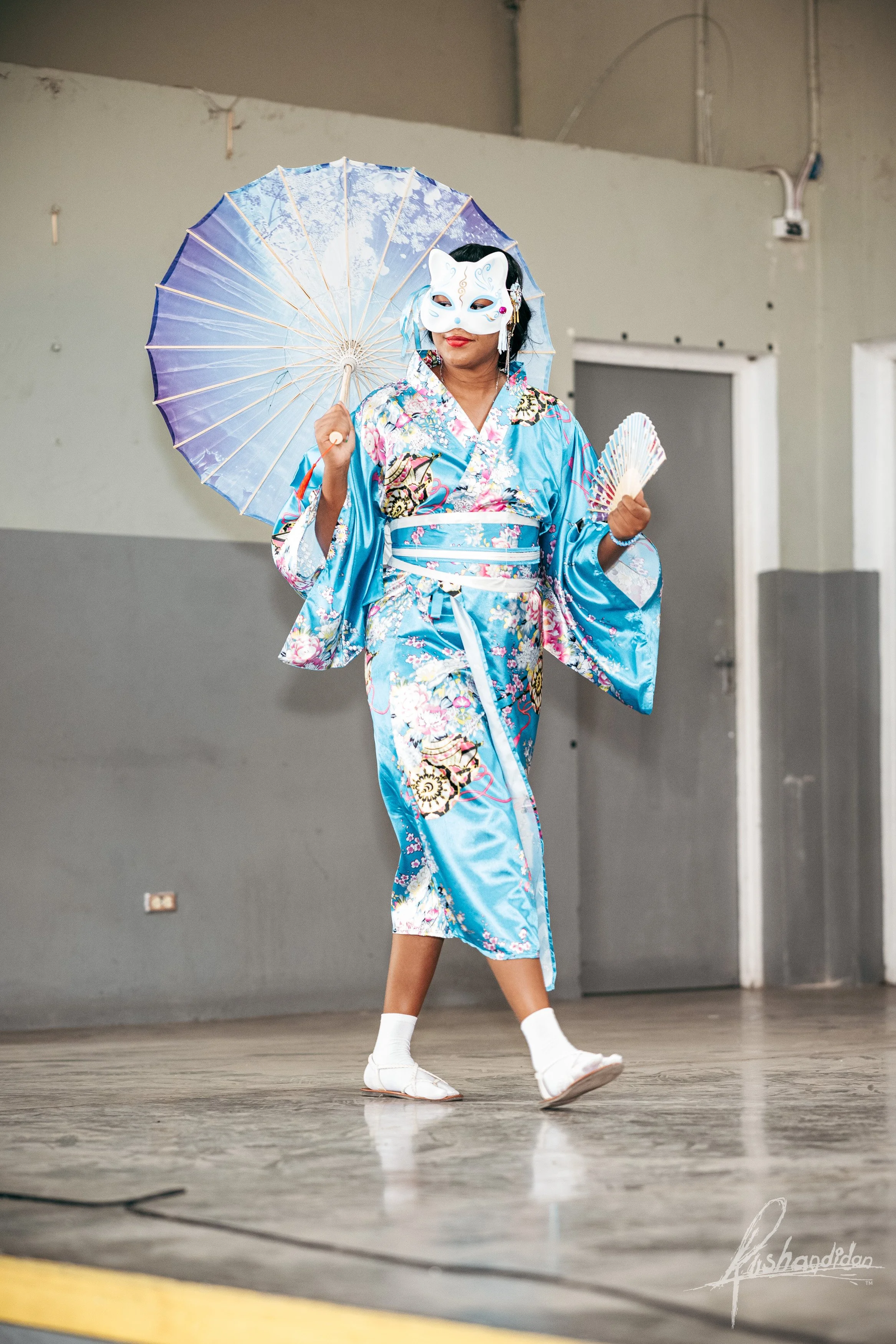 Person dressed in a blue traditional Japanese kimono holding an umbrella, wearing a white mask, and practicing a dance on an indoor stage.