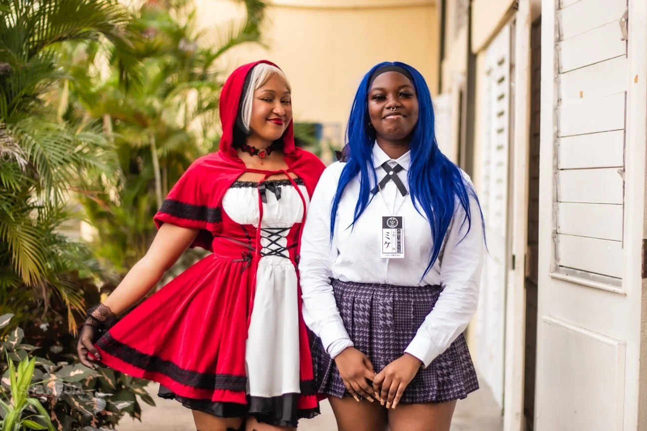 Two women standing outdoors near a white wooden building with greenery on the left. One woman is dressed as Little Red Riding Hood, wearing a red hooded cape and matching dress. The other woman has long, bright blue hair, a white shirt, a plaid skirt