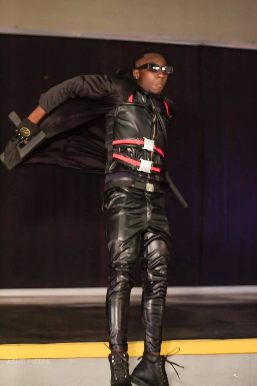 Person dressed in black leather outfit with red accents, wearing sunglasses, and holding a prop gun, standing on a stage with a dark background.