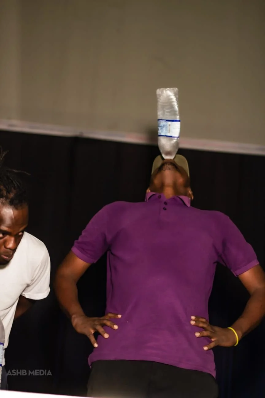 Person balancing a water bottle on their forehead while leaning back, with another person to the side, in an indoor setting.