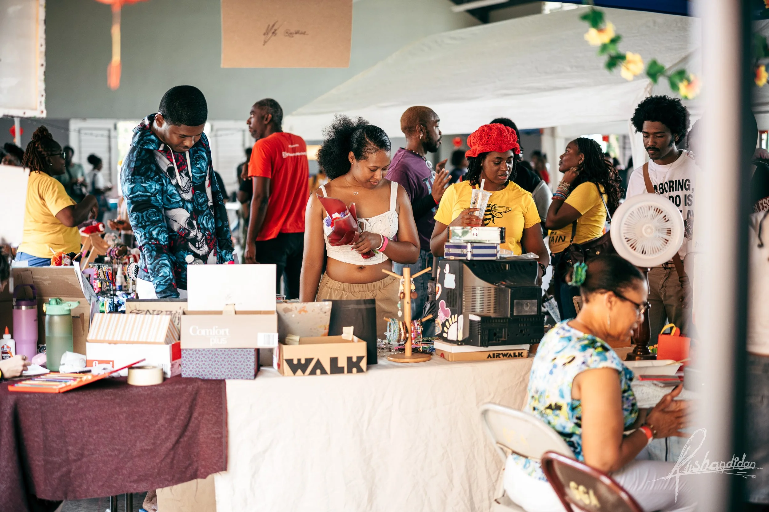 People shopping at an indoor market stall with various items on display, including boxes, a coffee machine, and decorative accessories. A woman seated at a table reads a book, while others browse and converse nearby.