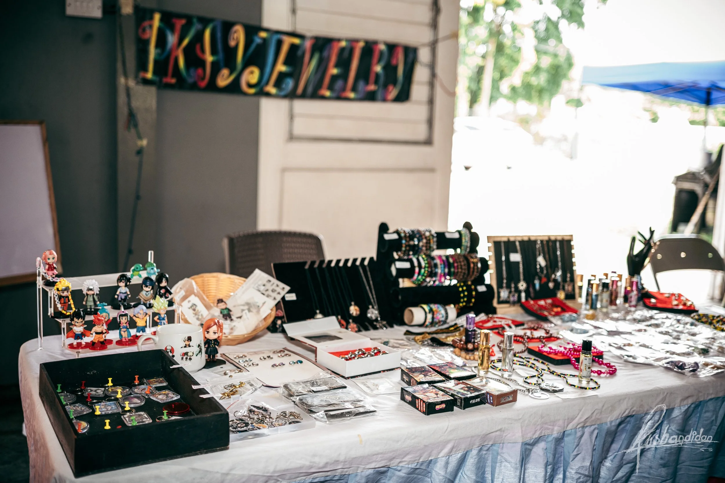 Table with assorted jewelry, toys, and collectibles displayed for sale at a market stall, with a colorful 'Happy Halloween' banner hanging in the background.