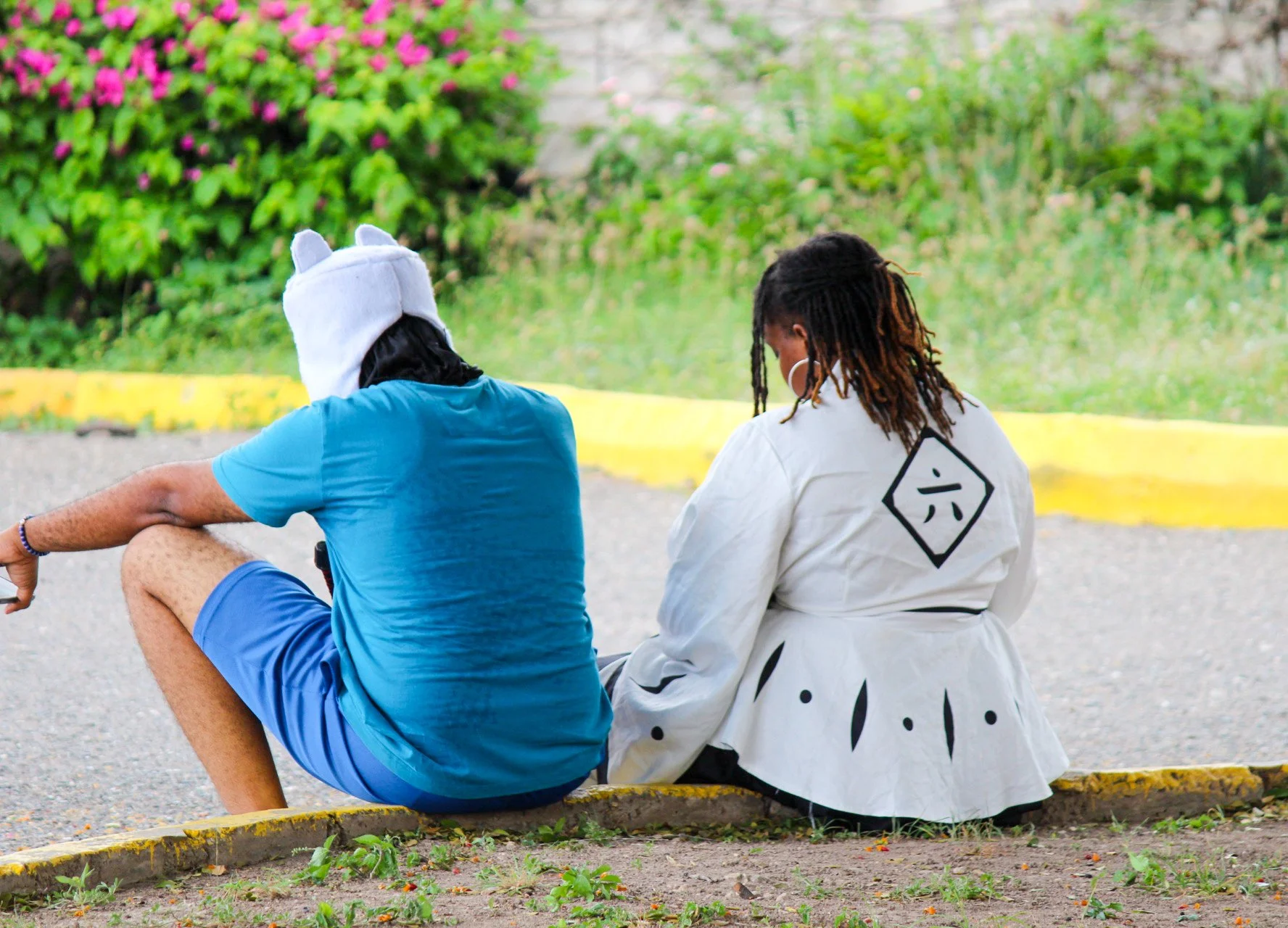 Two people sitting on the curbside outdoors near a garden with green bushes and pink flowers; one wears a blue t-shirt, shorts, and a hat with animal ears, the other wears a white jacket with a black design on the back and black pants.