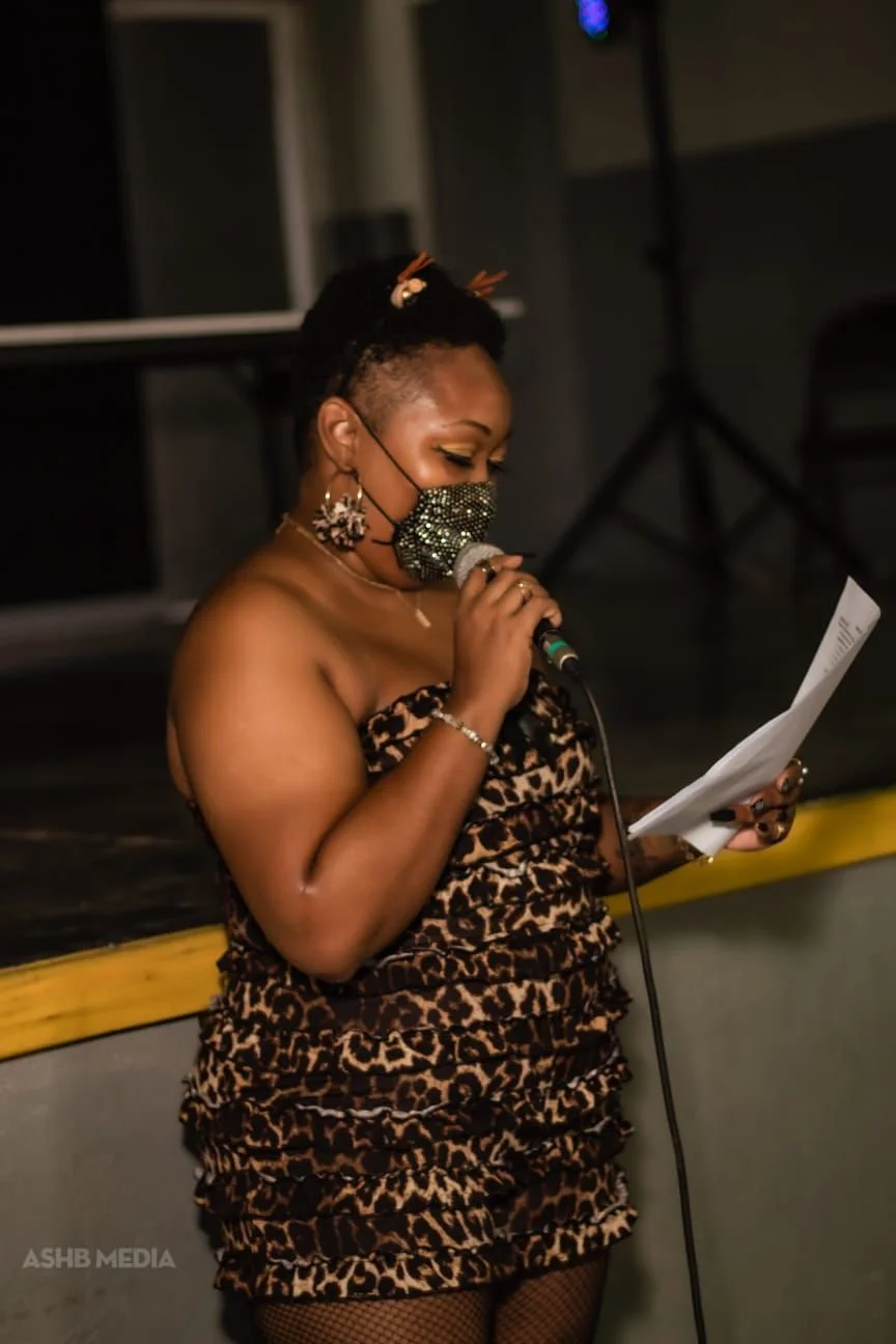 A woman with styled hair and a leopard print dress, wearing a sparkly mask, holding a microphone and reading from a sheet of paper.