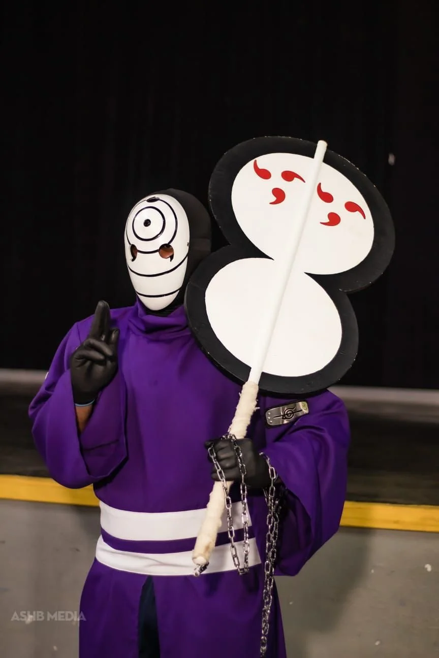 Person dressed in purple costume with a chain, wearing a mask with a spiral design, holding a fan that resembles a yin-yang symbol with red marks, on a dark background.