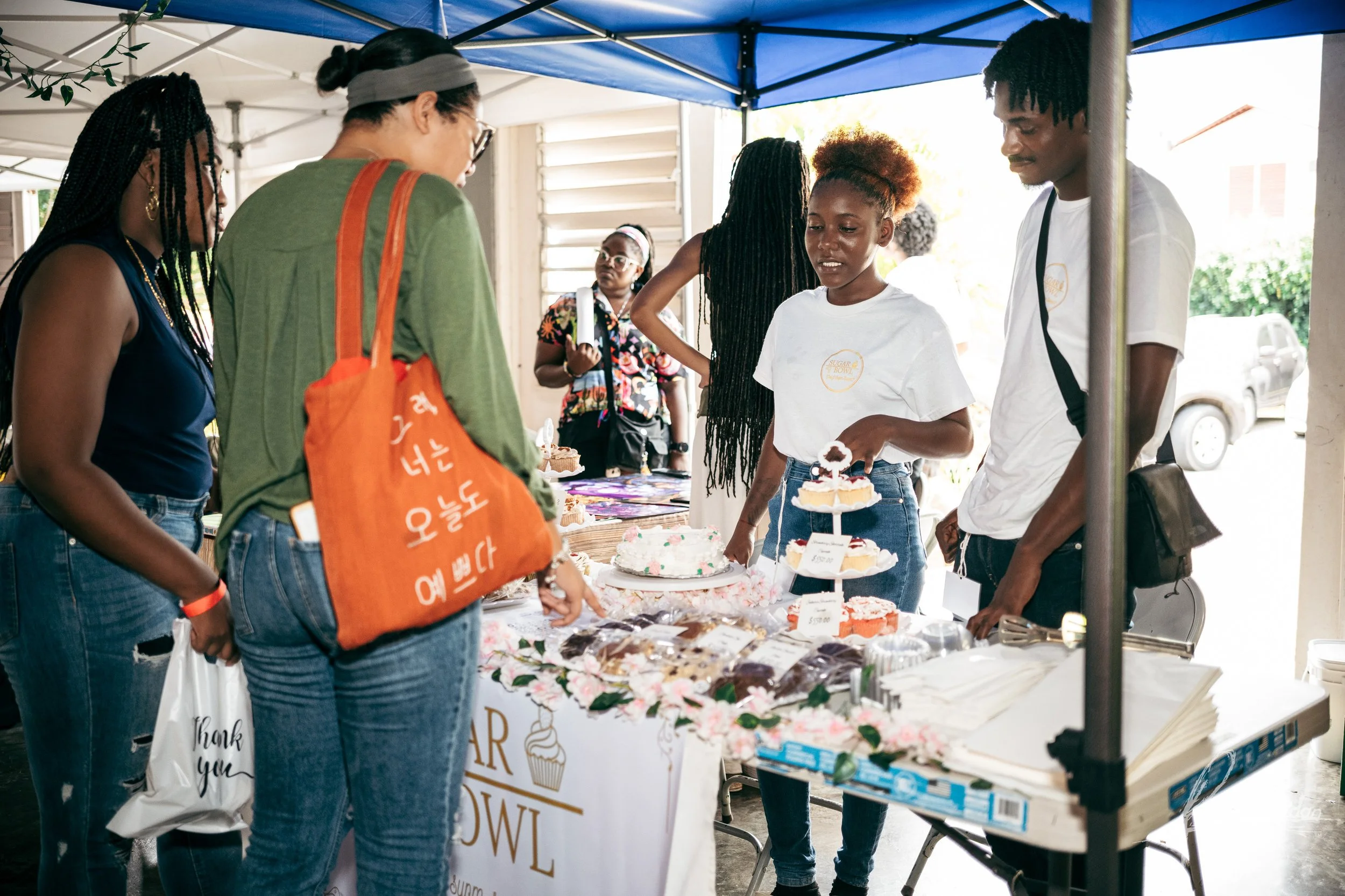 A group of people are gathered around a table with baked goods at an outdoor market or fair. There are cakes and desserts on display, and a woman behind the table is serving or selling them. The setting is under a canopy tent.