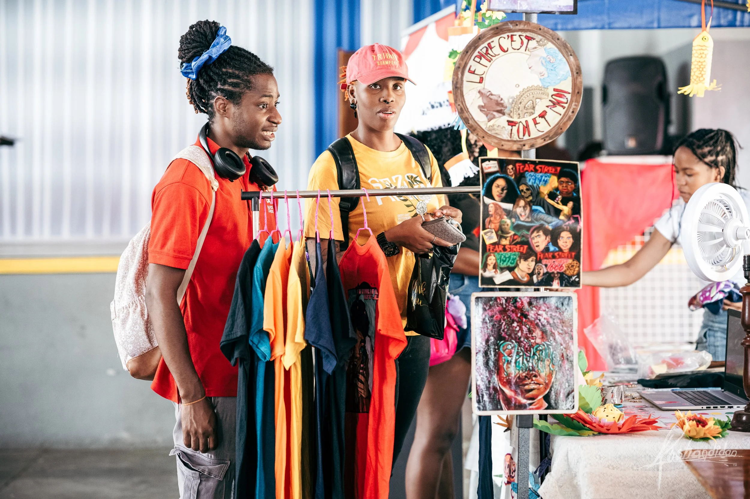 Two women shopping at a merchandise stand selling colorful T-shirts, artwork, and prints at an indoor market or fair.