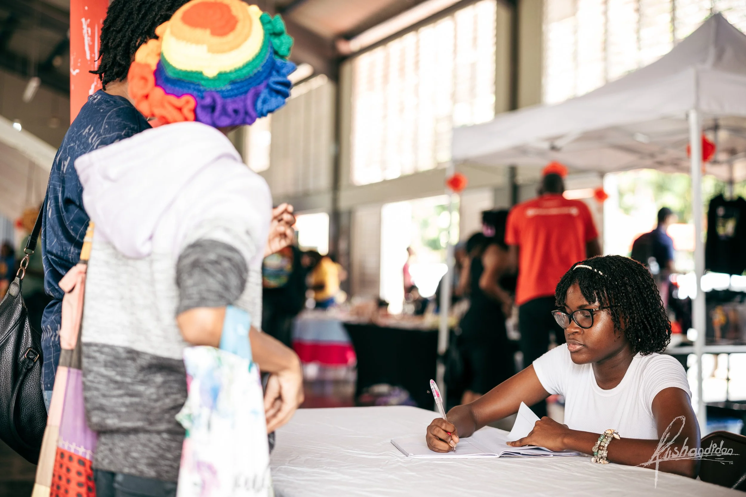 A woman with glasses and curly black hair, wearing a white t-shirt, sits at a table and writes in a notebook. She is at an indoor event, with a man wearing a rainbow-colored knit hat and a woman with a gray hoodie and backpack standing in front of he