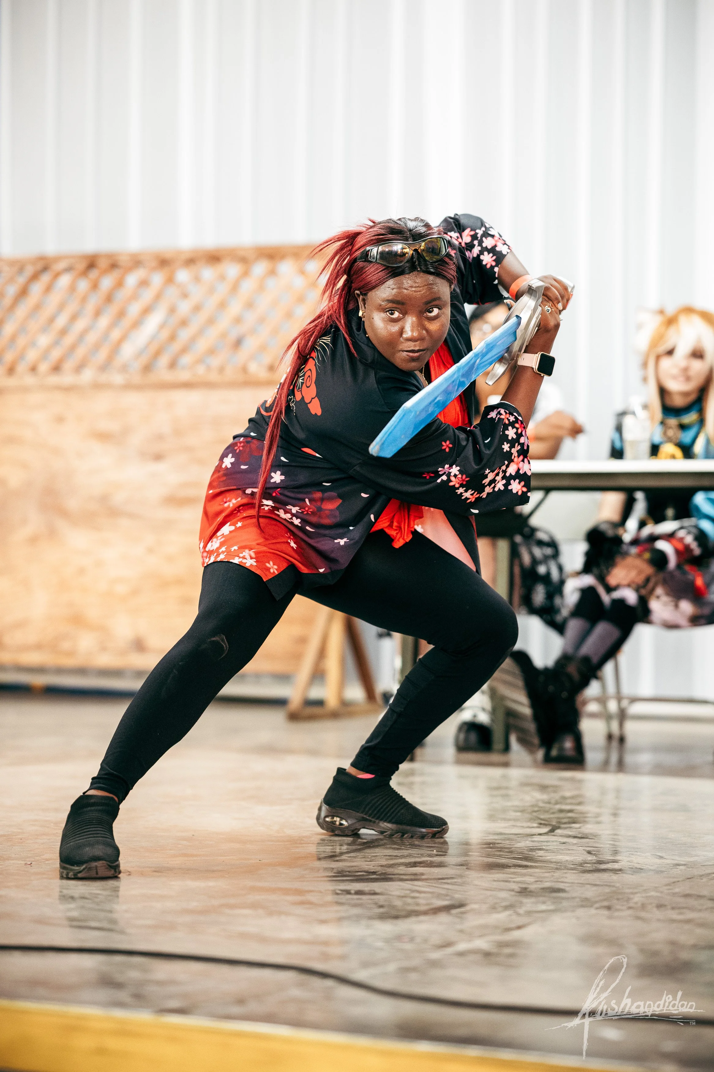 A woman in black and red sportswear, with red hair and sunglasses on her head, is in a low stance holding a blue sword with both hands during a martial arts demonstration or training session in an indoor space with a wooden floor and white walls. Two