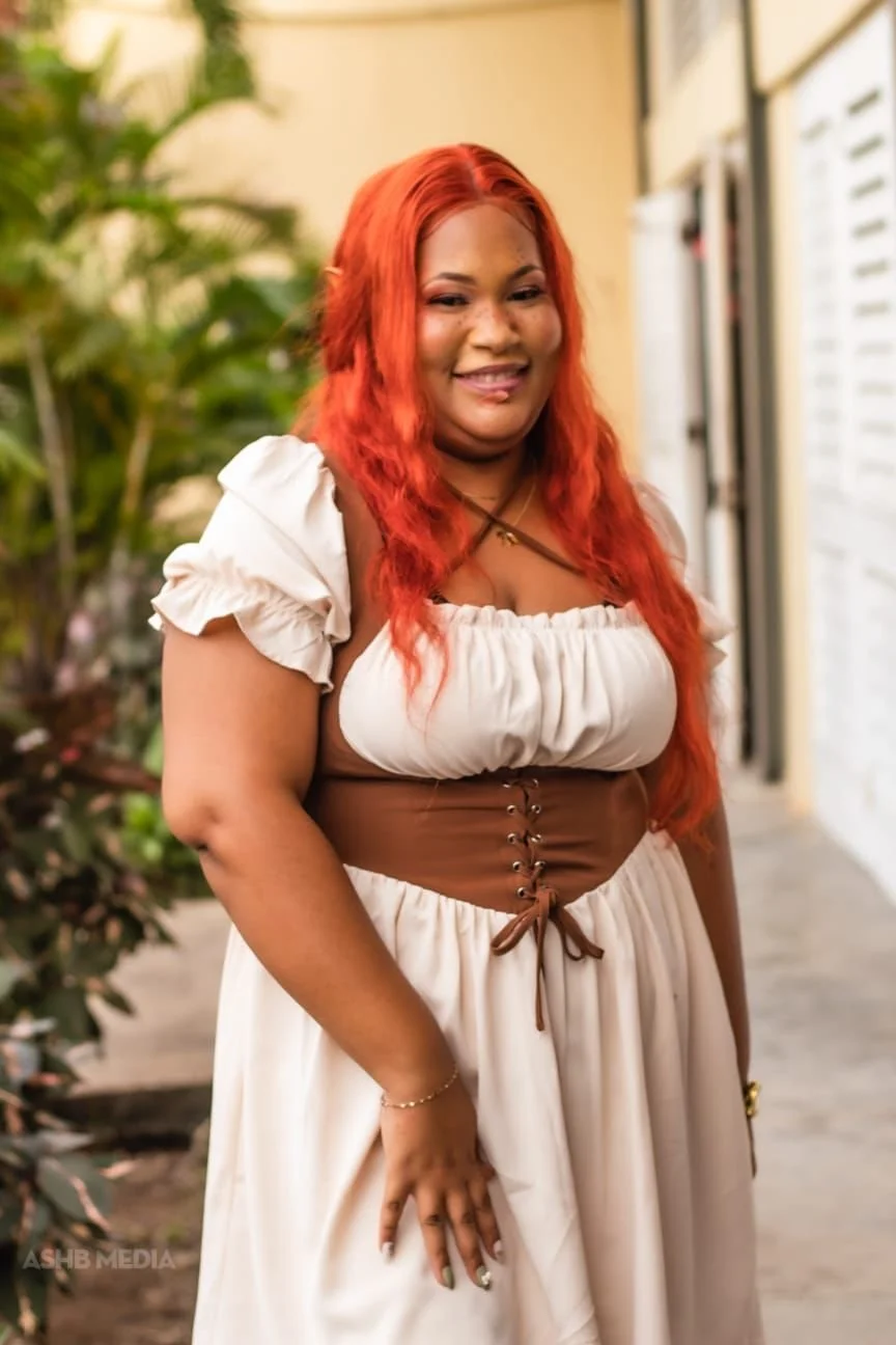 A woman with long, wavy red hair smiling, wearing a cream-colored dress with puffed sleeves and a brown corset-style belt, standing outdoors with greenery in the background.