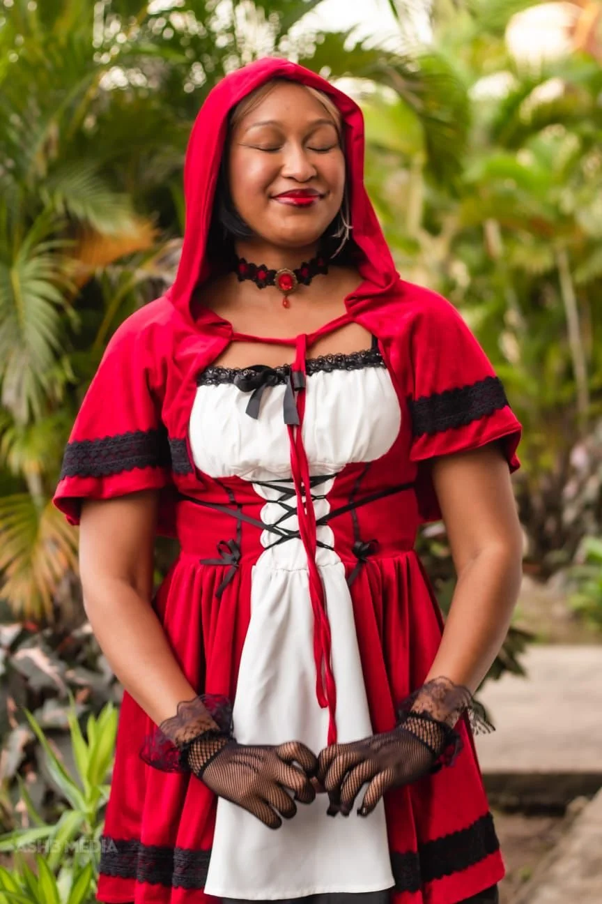 A woman dressed in a costume inspired by Little Red Riding Hood, with a red hooded cape, black lace accents, black gloves, and a Gothic-style choker, standing outdoors among lush green plants with her eyes closed and smiling.