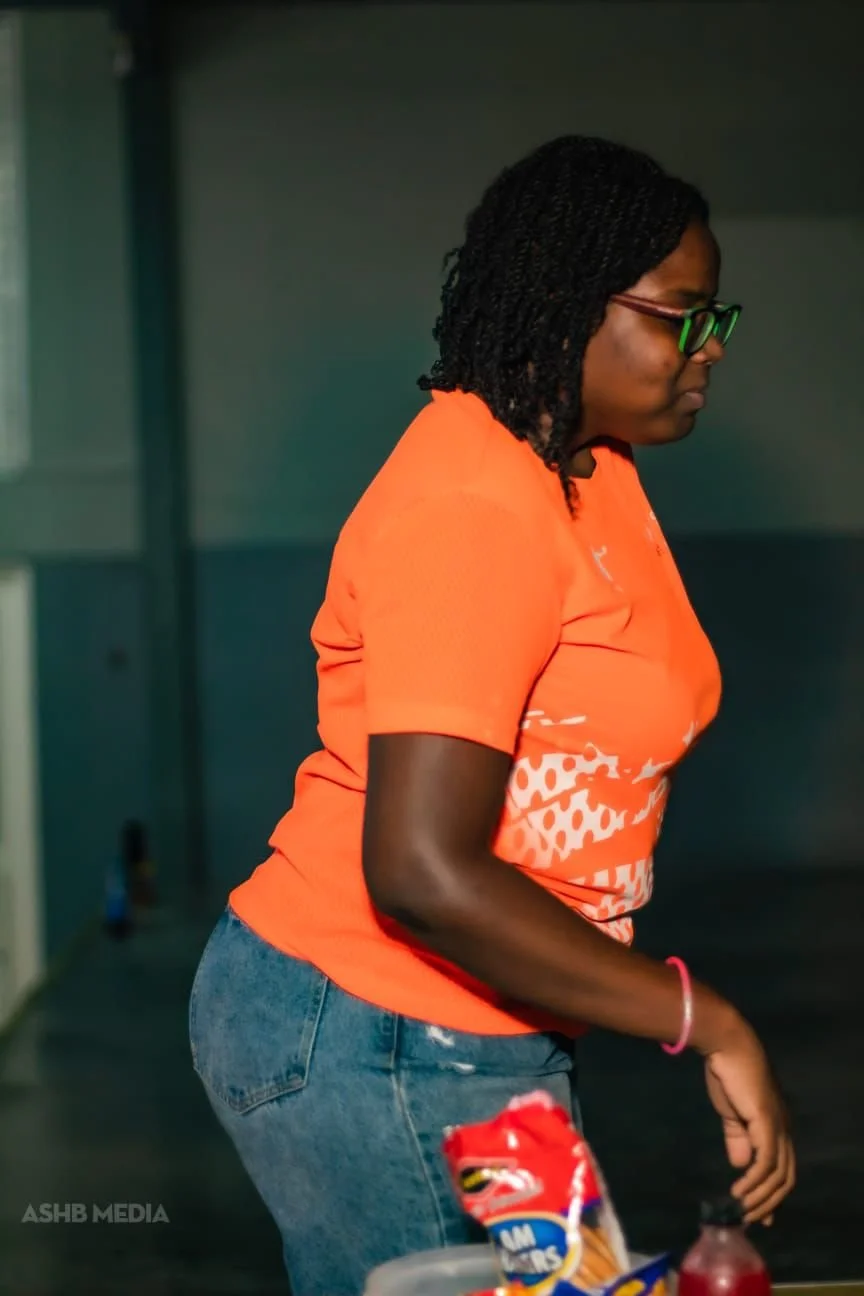 A woman with glasses, wearing an orange shirt and jeans, standing indoors.