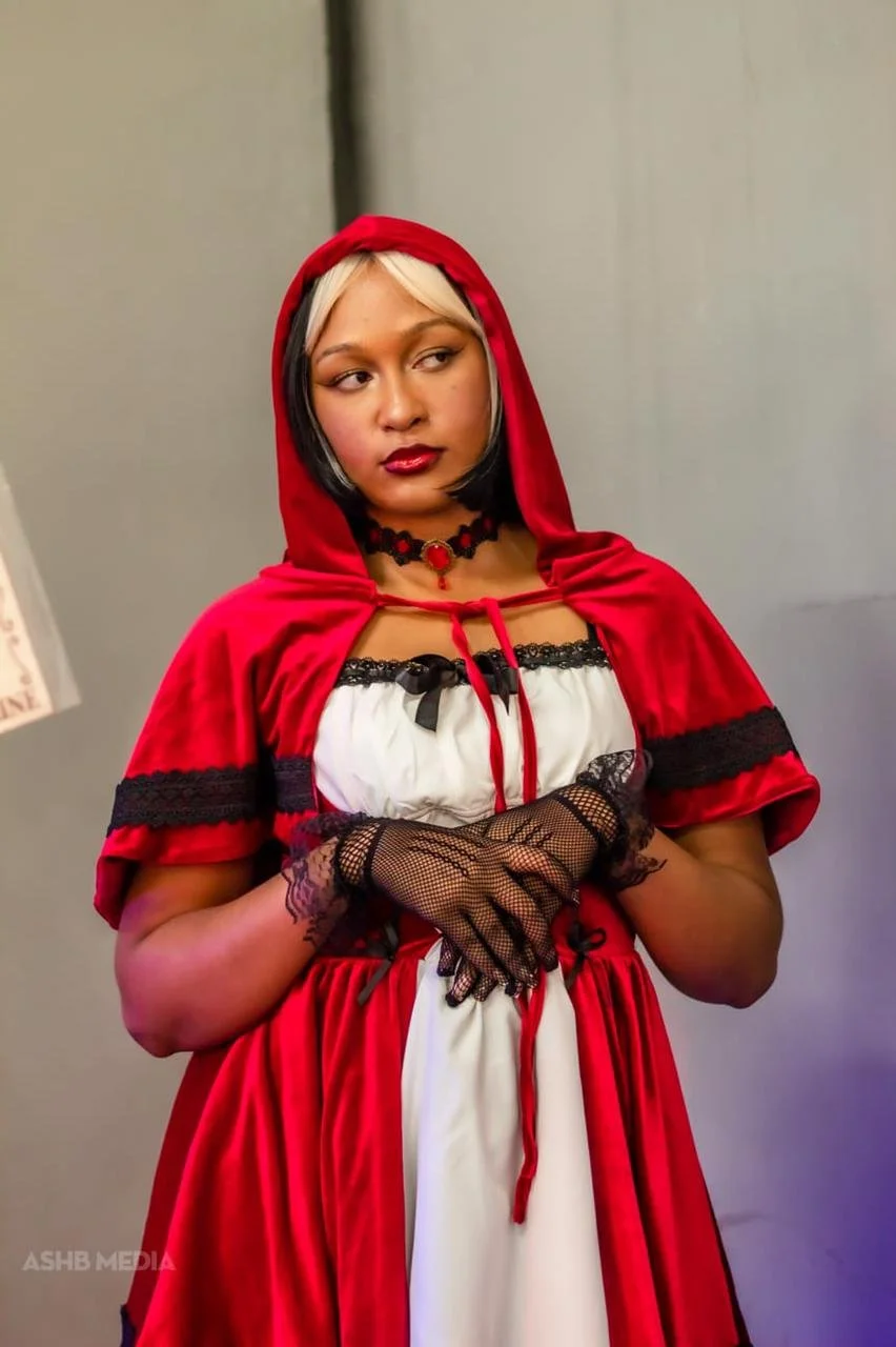 A woman dressed in a red hooded cape, wearing black lace gloves, and a black choker, looking to the side with a serious expression.