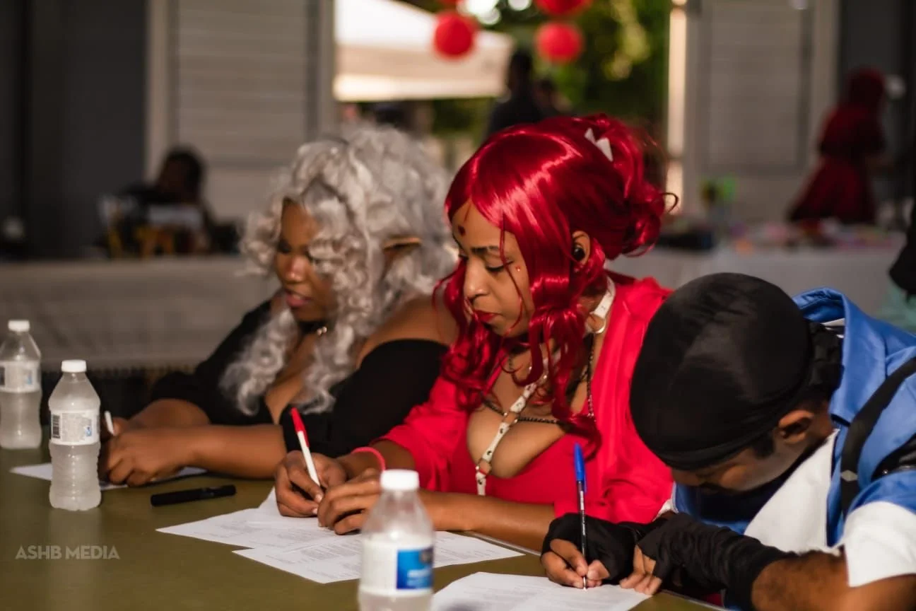 Three people in colorful costumes sit at a table, writing on papers with water bottles and pens in front of them. One woman has long red hair and a revealing red outfit, another has curly gray hair, and the third wears a black and blue costume with a