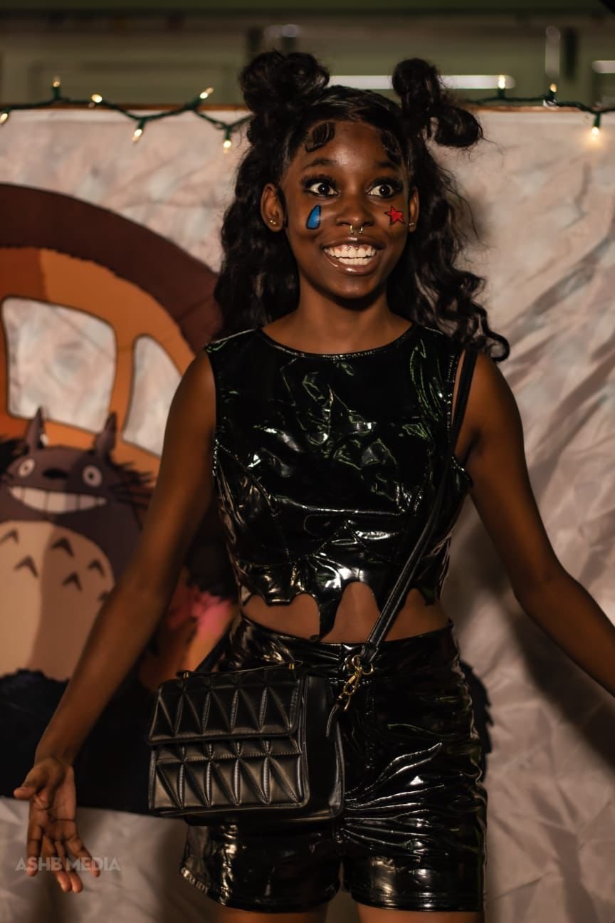 A young woman with dark curly hair styled into two buns, wearing face stickers of a blue teardrop and a red star, smiling in front of a backdrop featuring a cartoon creature and string lights, dressed in a black shiny outfit with matching shorts and 