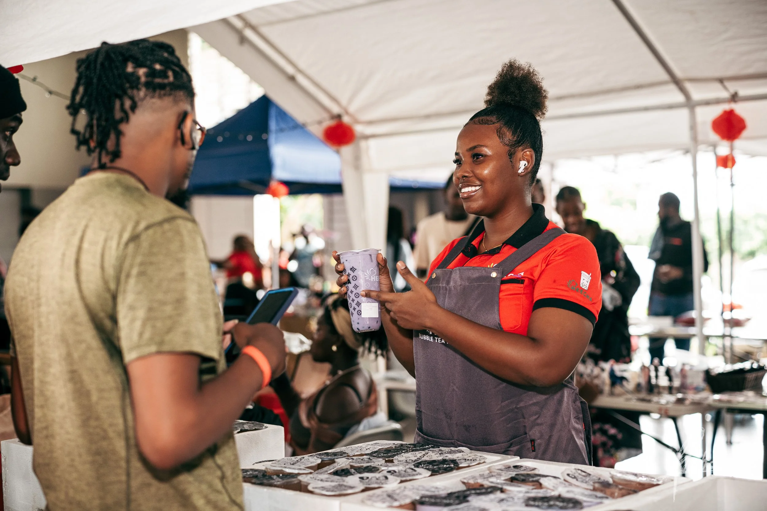 A smiling woman selling jewelry at an outdoor market interacts with a customer, who is looking at her phone. The woman is wearing a red and black uniform with a gray apron. The market is under a tent with other vendors and shoppers in the background.