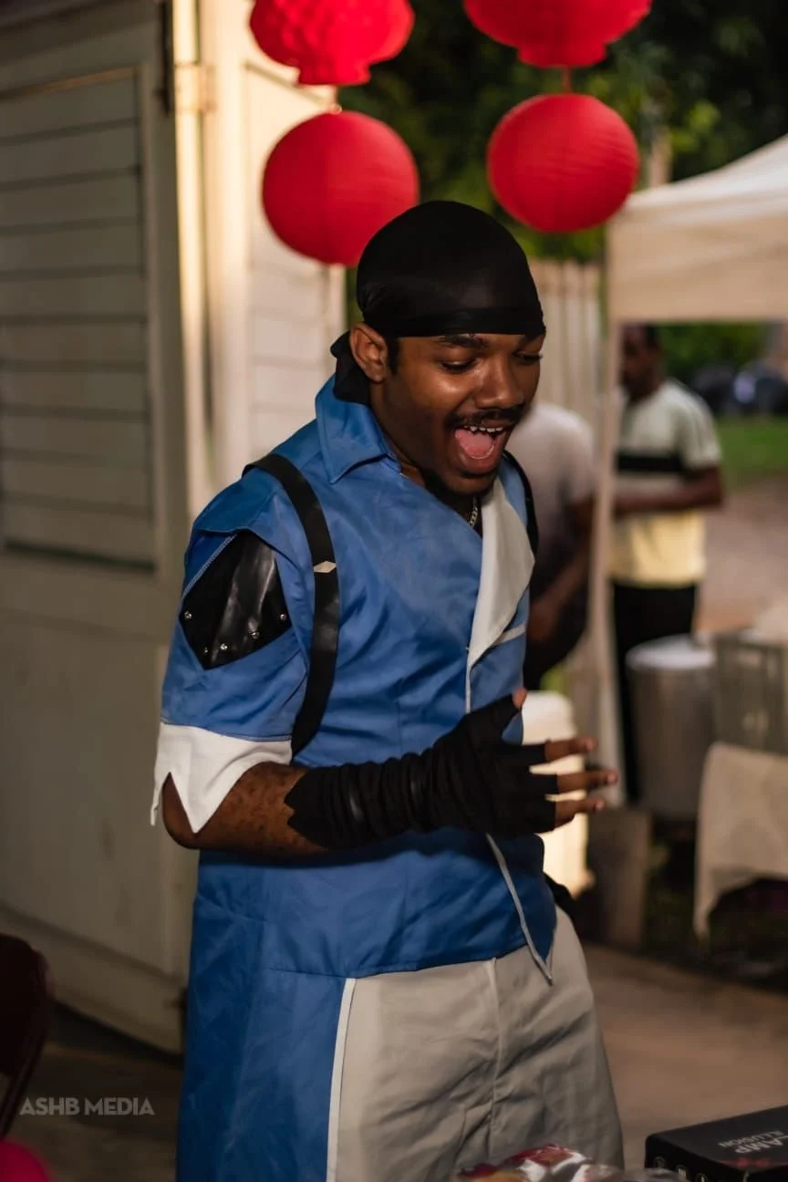 A young man wearing a blue and white jacket, a black durag, and black gloves, smiling and looking down at a table at an outdoor event with red lanterns hanging overhead.