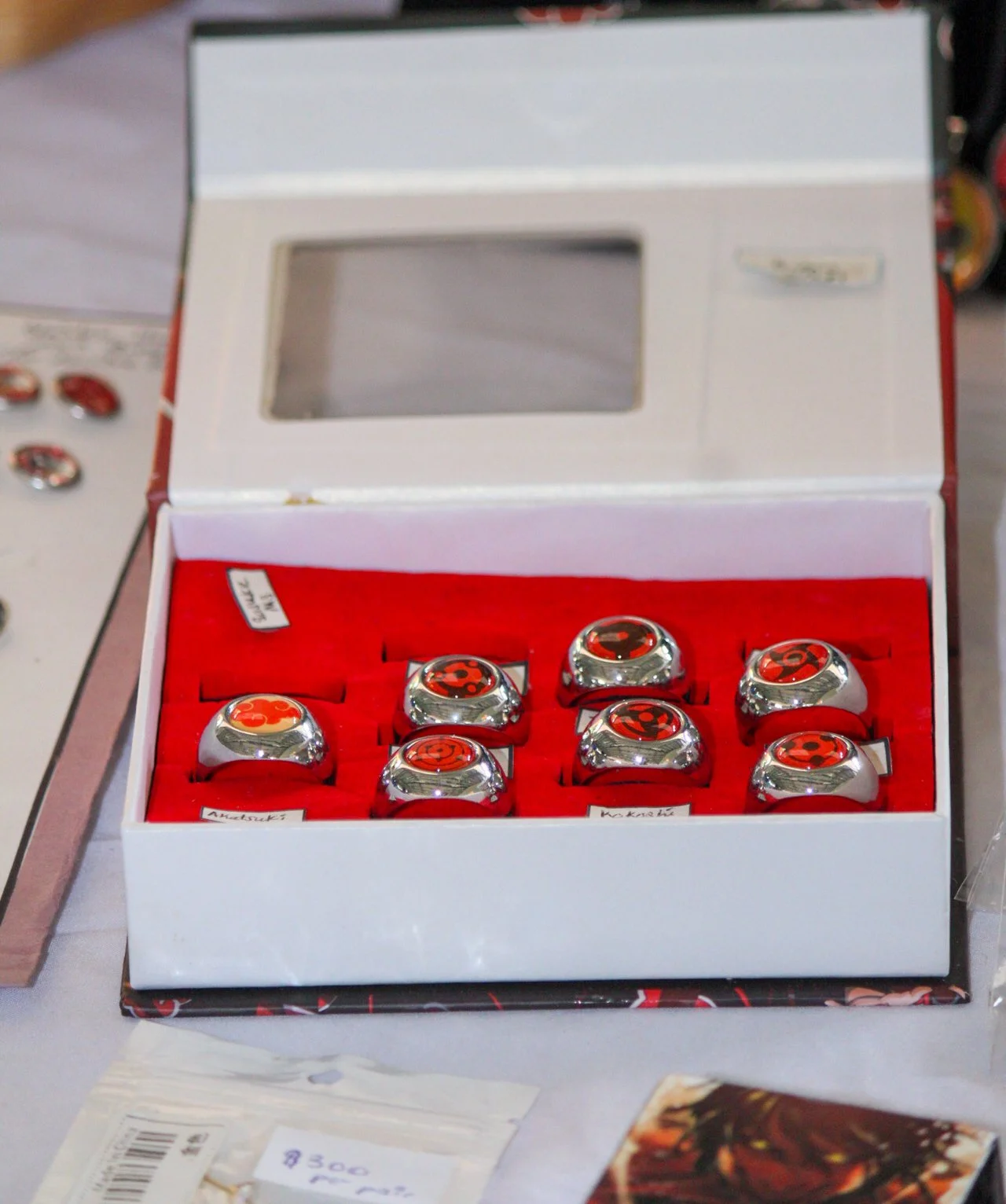 A white jewelry box with an open lid displaying six silver rings, each with a red and black clutch design, on a red velvet interior.