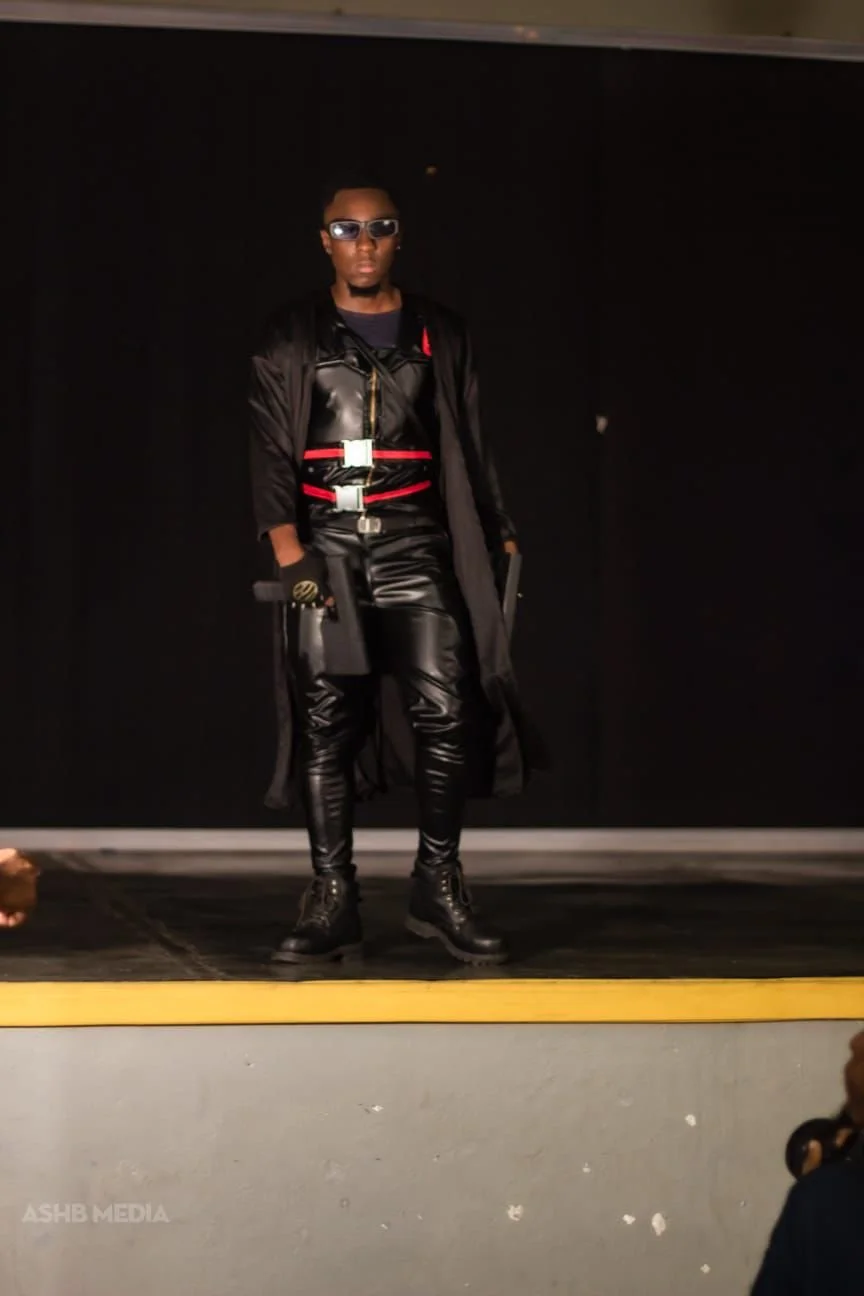 A man dressed in black leather outfit with red and white accents, wearing sunglasses and black boots, standing on a stage with a black background.