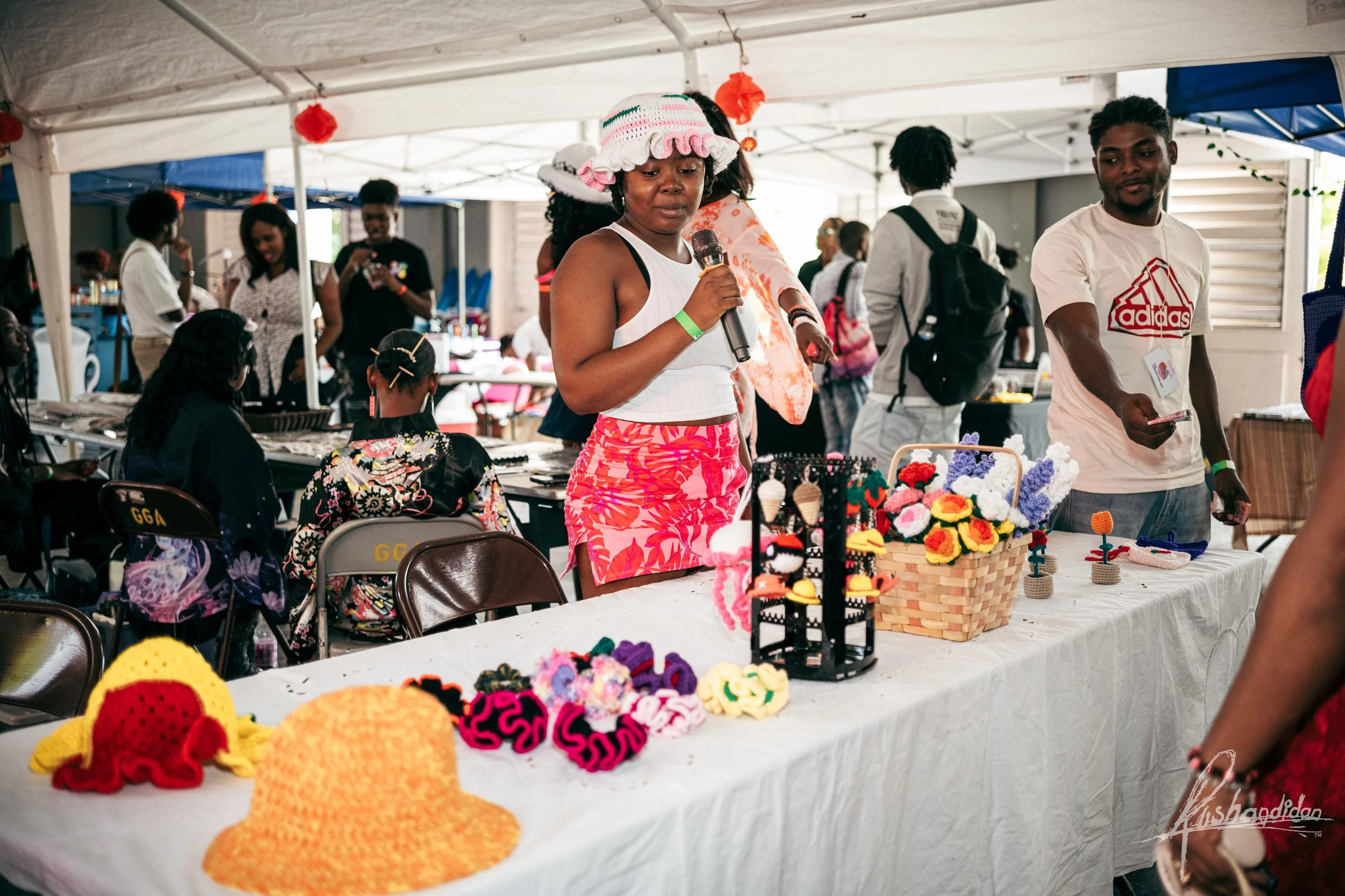 A woman at a craft fair displays handmade crocheted hats and accessories on a table, with other people browsing and selling items in the background under a white tent.