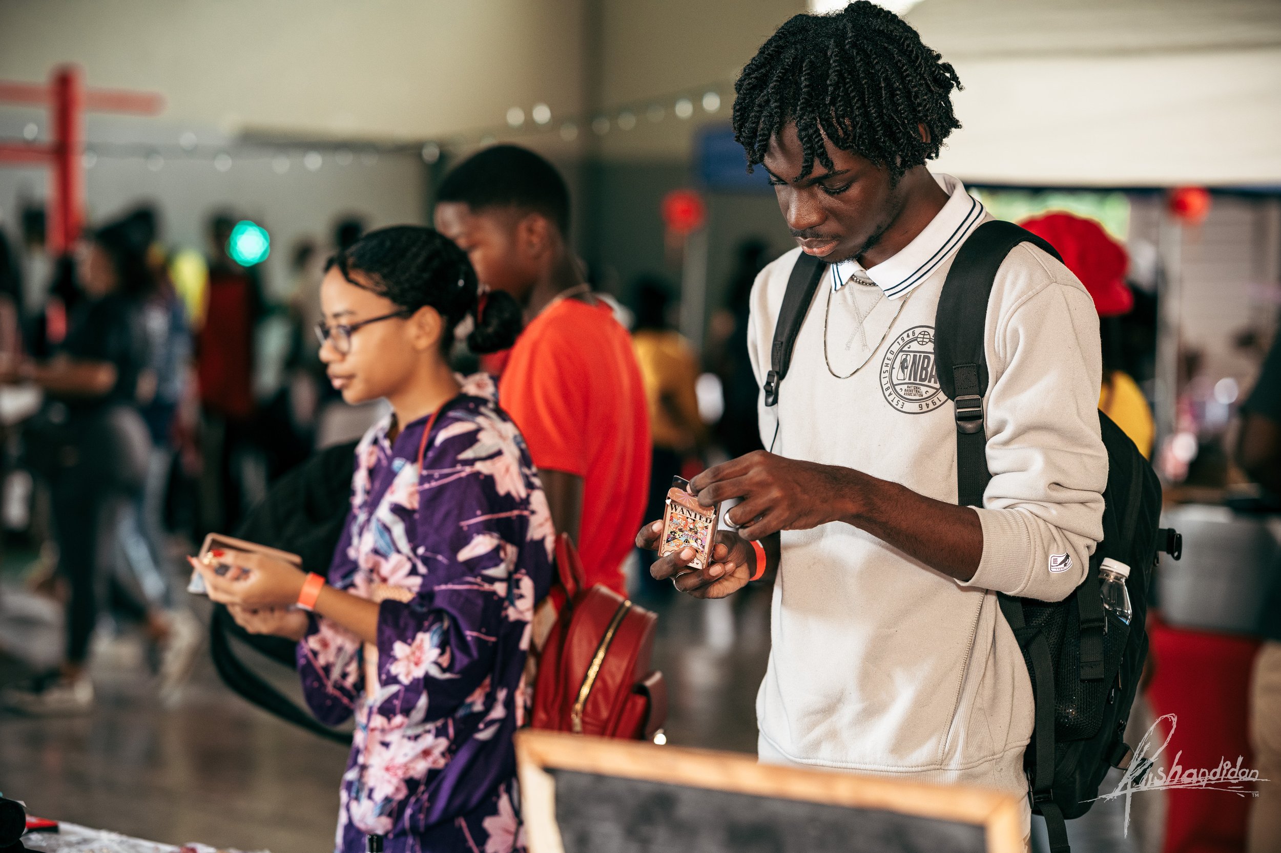 A group of young people standing in line indoors, with some reading cards or items in their hands, and one person looking at a small item.