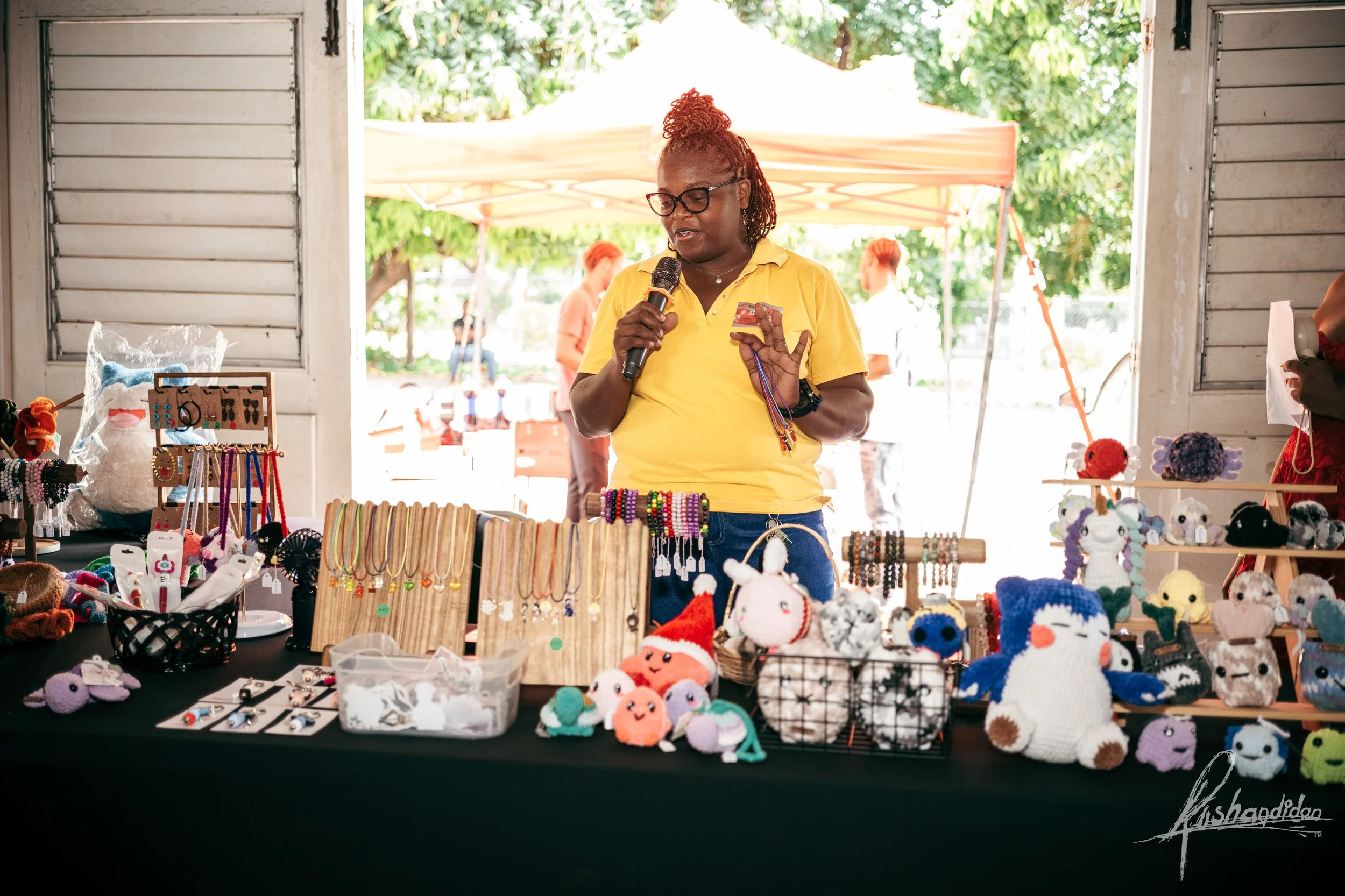 Woman in yellow shirt selling handmade jewelry and plush toys at an outdoor craft market stall with various colorful items displayed on black tablecloth and a canopy with trees and people in the background.