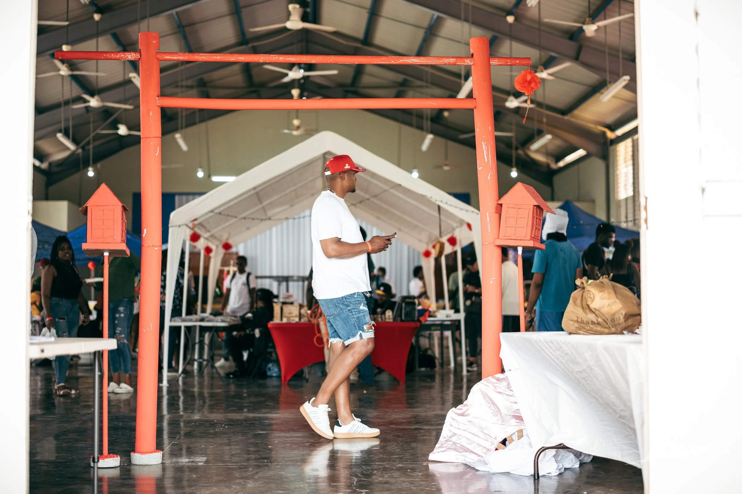 Man in white shirt, denim shorts, red-cap, and sneakers standing indoors with event booths and people in the background.