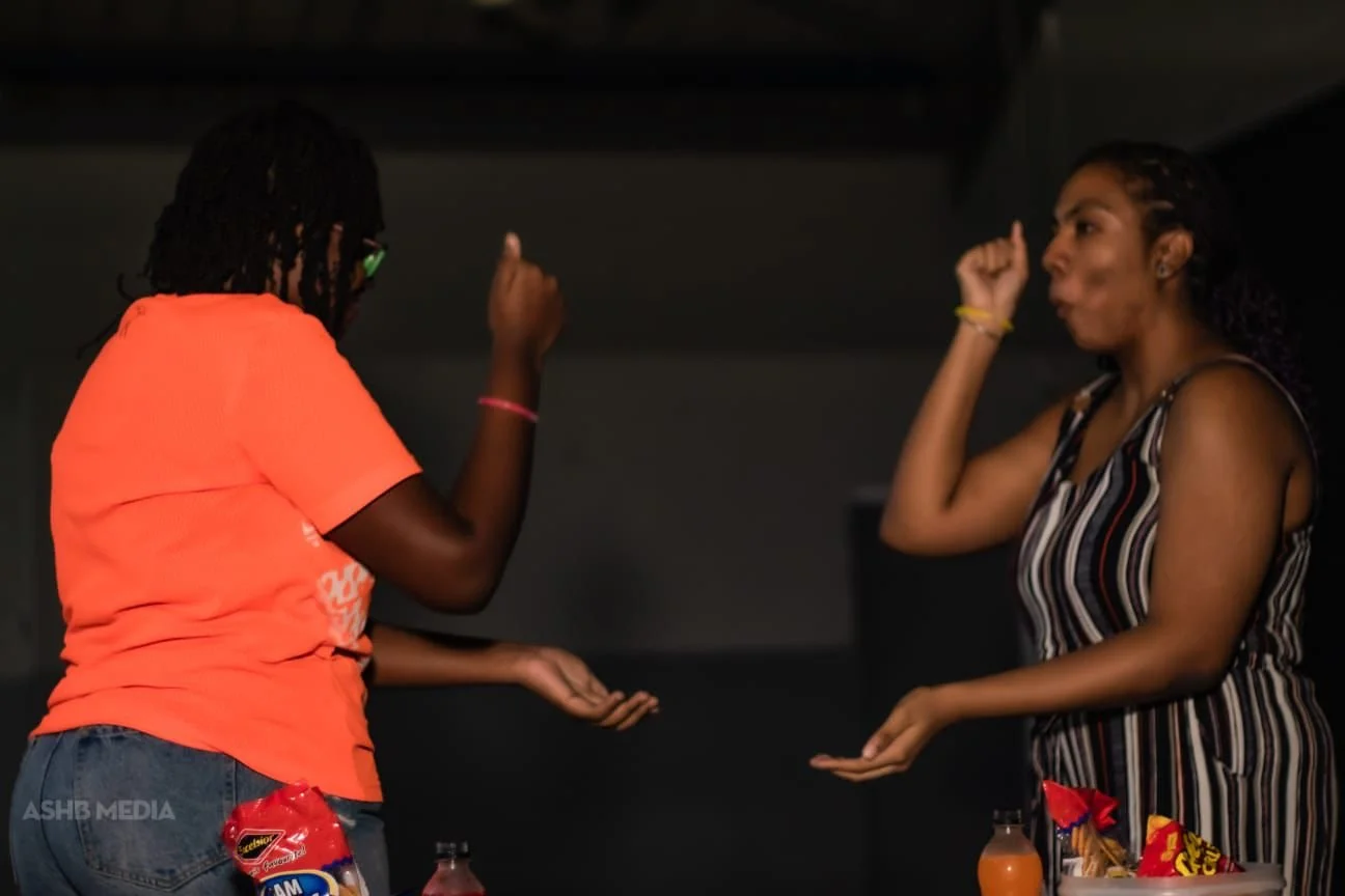 Two people appear to be engaged in a playful or animated conversation, facing each other with one person's fists raised and the other's hands open in a gesture, in a dimly lit room.