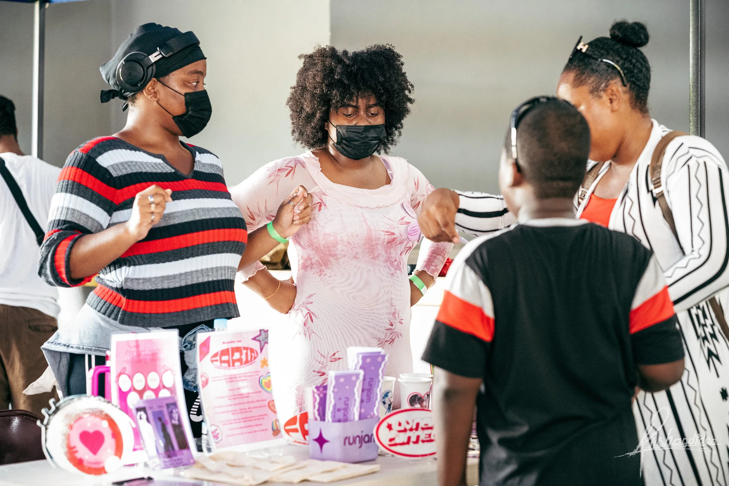 Four women, two wearing masks, are gathered around a table with pink and purple promotional items, engaging in conversation at an indoor event.