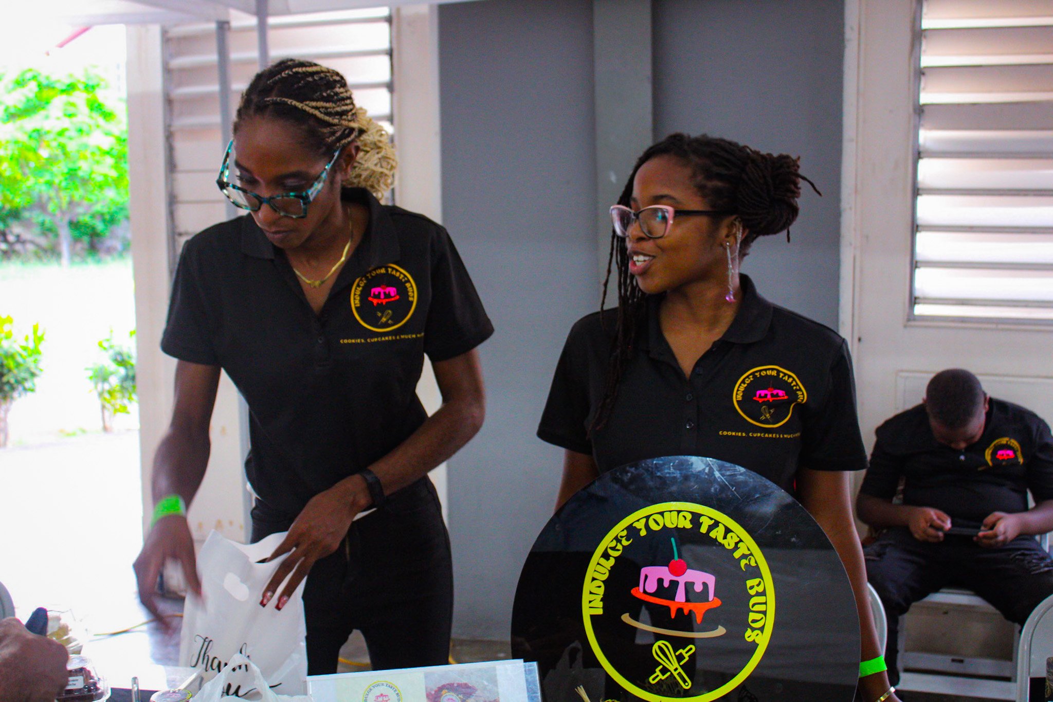 Two women wearing black shirts with a logo that reads 'Indulge Your Taste Buds' are standing at a table, with one woman looking down at items and the other woman talking. A man in the background is sitting and looking at his phone.