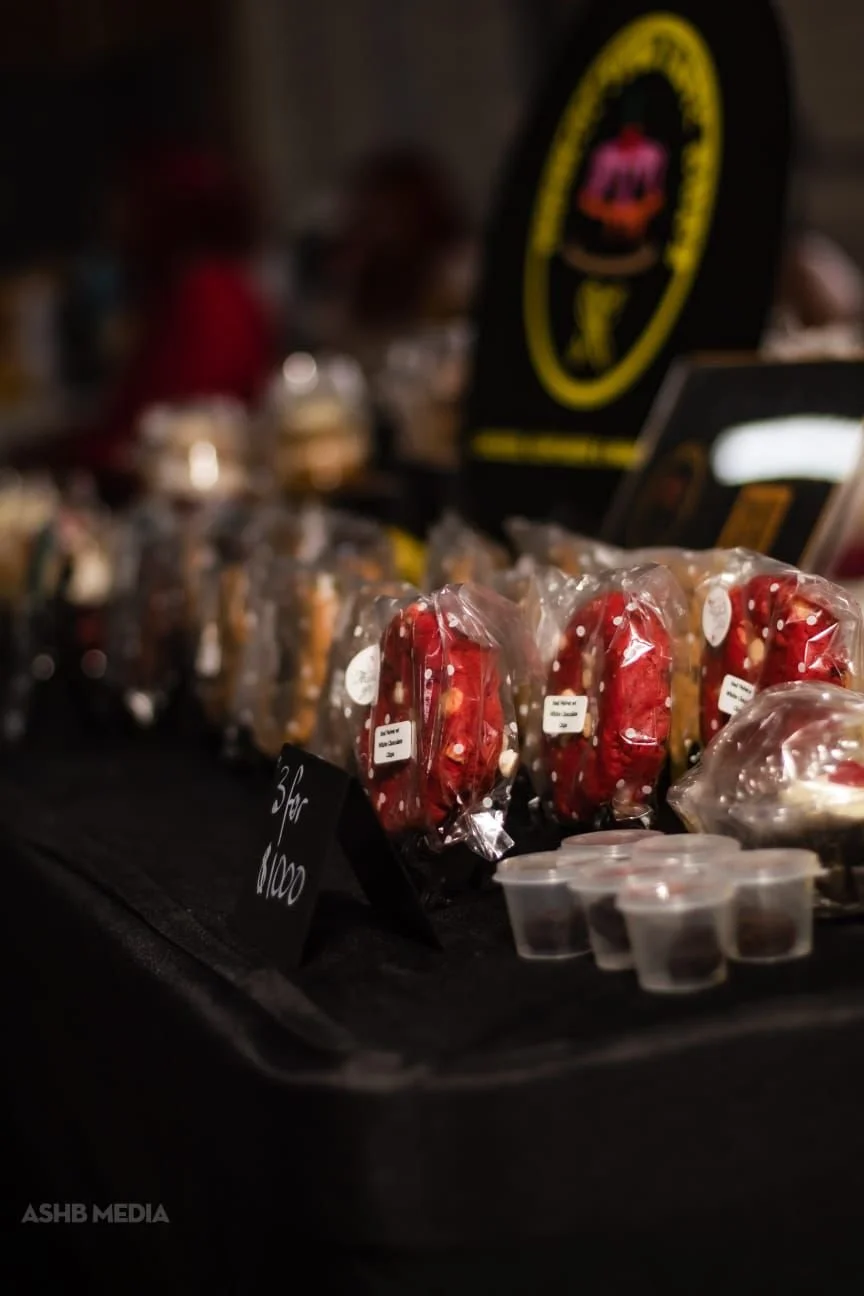 Candy apples wrapped in plastic on display at a market or store, with small containers of toppings or dips in front and a black sign with white writing.