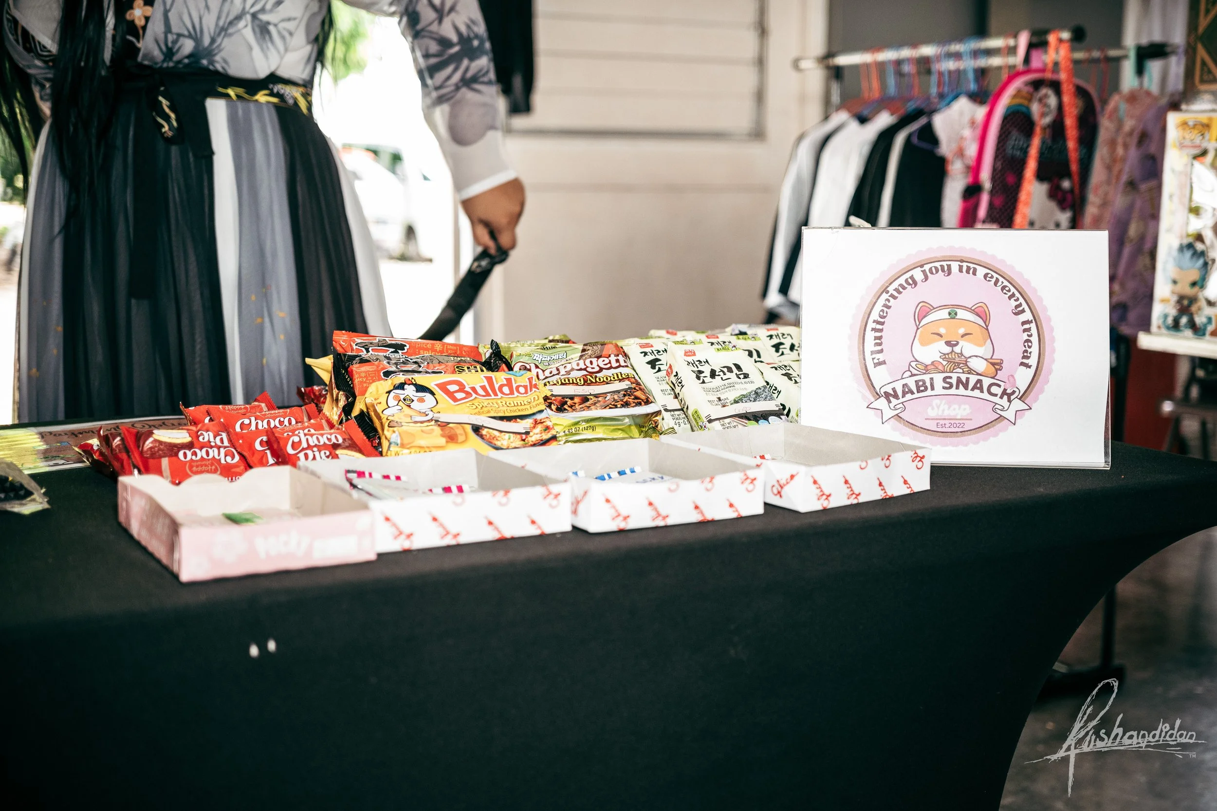 Display table with assorted snack packages, including instant noodles and cookie bags, set up at an outdoor market stall. A sign with a cartoon cat and the text "NABI SNACK Shop" is visible on the right.