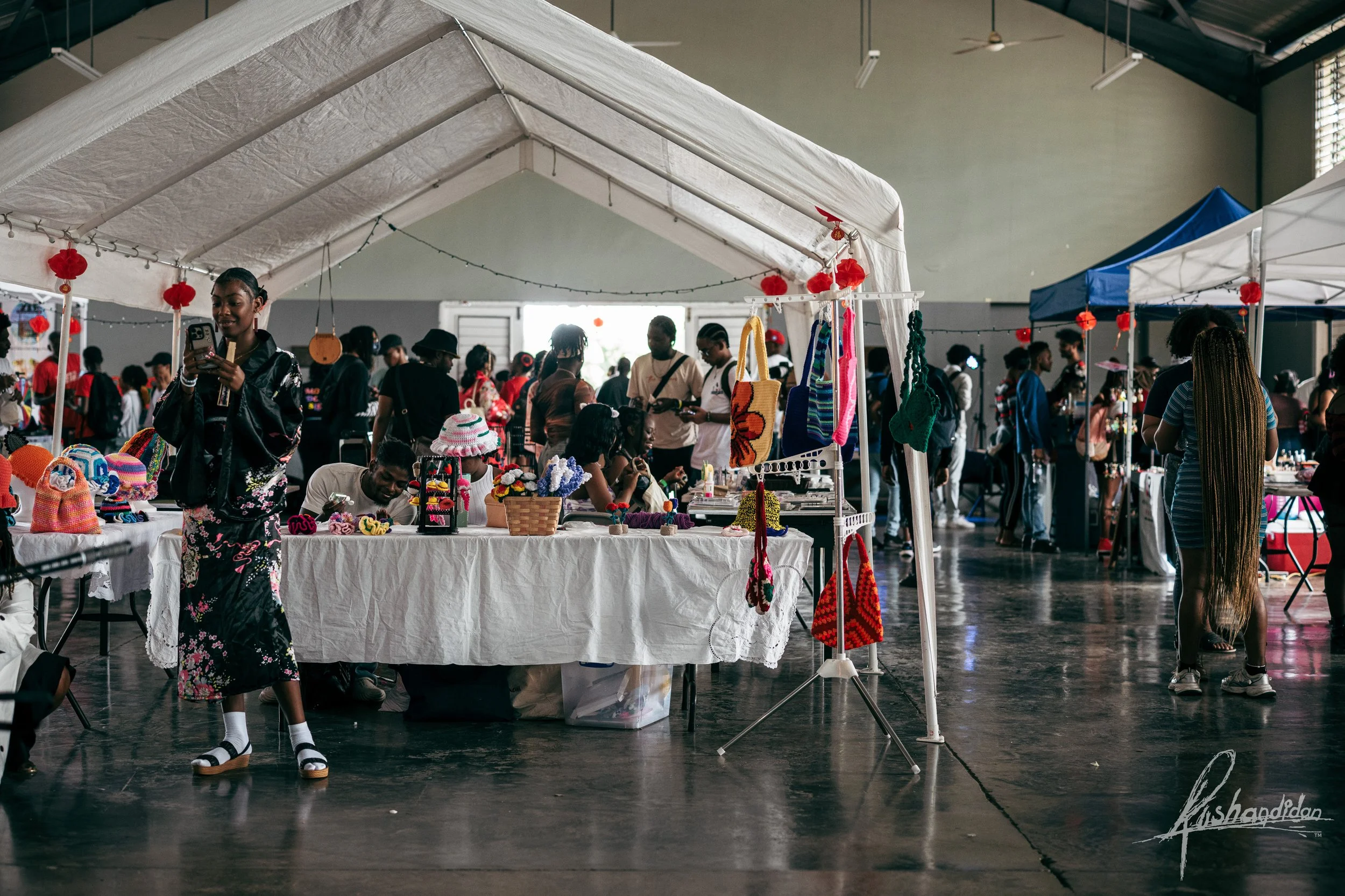 People shopping at an indoor craft market with various vendors selling handmade items such as hats, bags, and accessories, under a large white tent.
