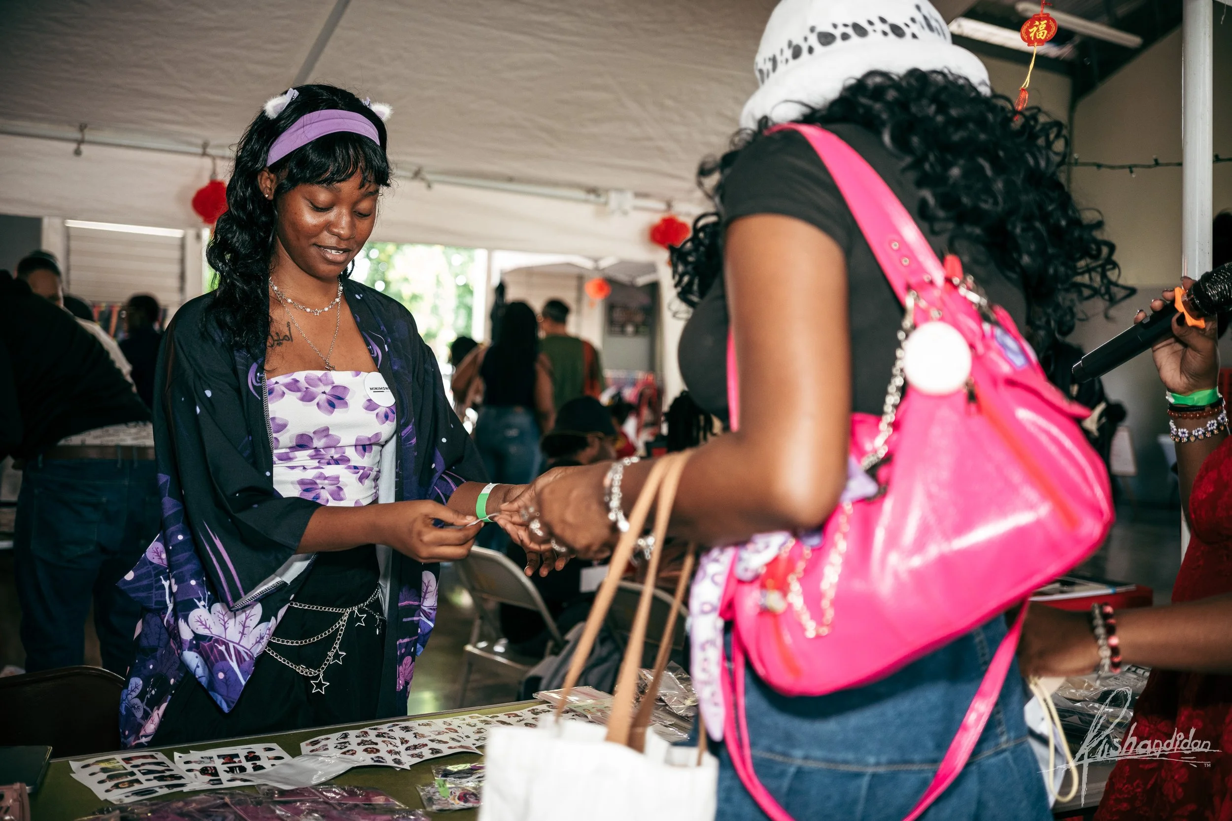 A woman with dark curly hair, wearing a black top and bright pink backpack, is purchasing jewelry from a vendor at a market stall. The vendor has long dark hair, wearing a purple and white floral top with a dark jacket. The vendor is handing a jewelr