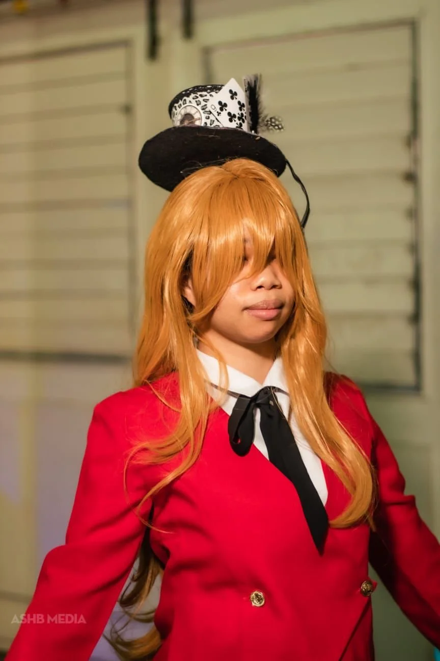 Person with long orange hair wearing a red jacket with gold buttons and a white shirt with a black ribbon, balancing a small black top hat with a feather and a deck of playing cards on their head.