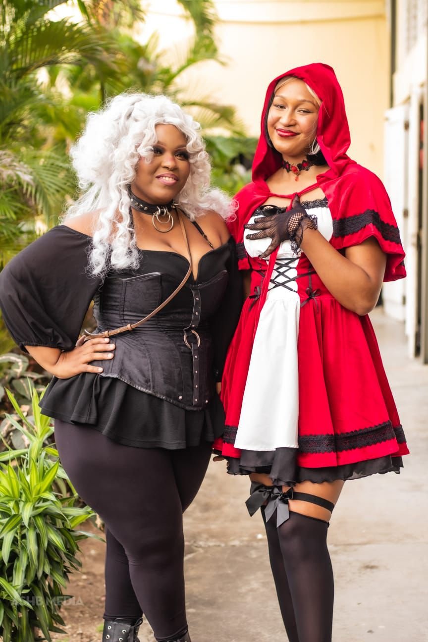 Two women dressed in costumes, one as Little Red Riding Hood and the other in a gothic black outfit, standing outdoors among green plants and smiling.