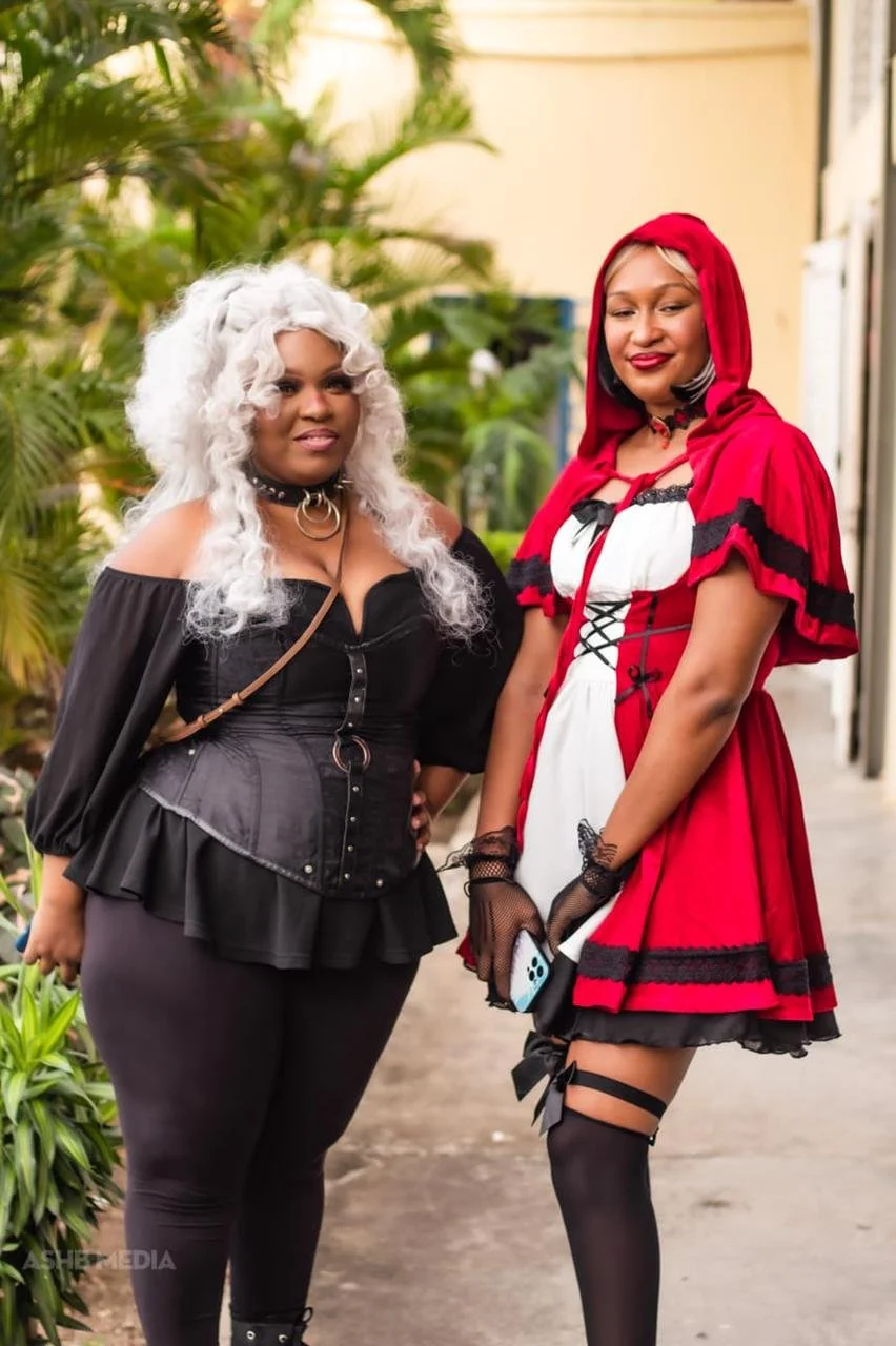 Two women dressed in costumes, one as Little Red Riding Hood and the other in Gothic attire, standing outdoors with greenery in the background.