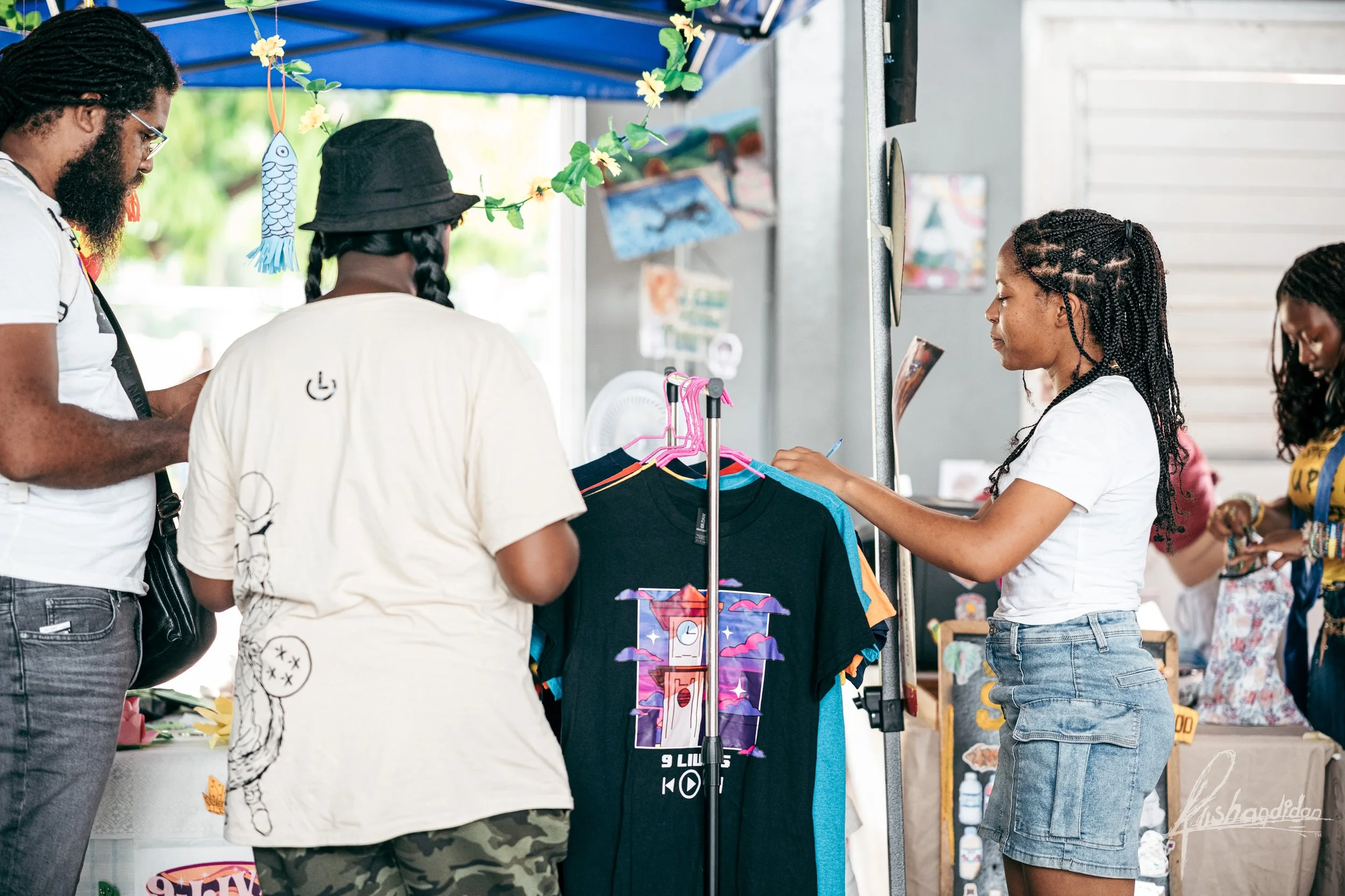 Three women and one man at an outdoor stall selling clothing, with a rack of T-shirts and decorative hanging fish and flowers.