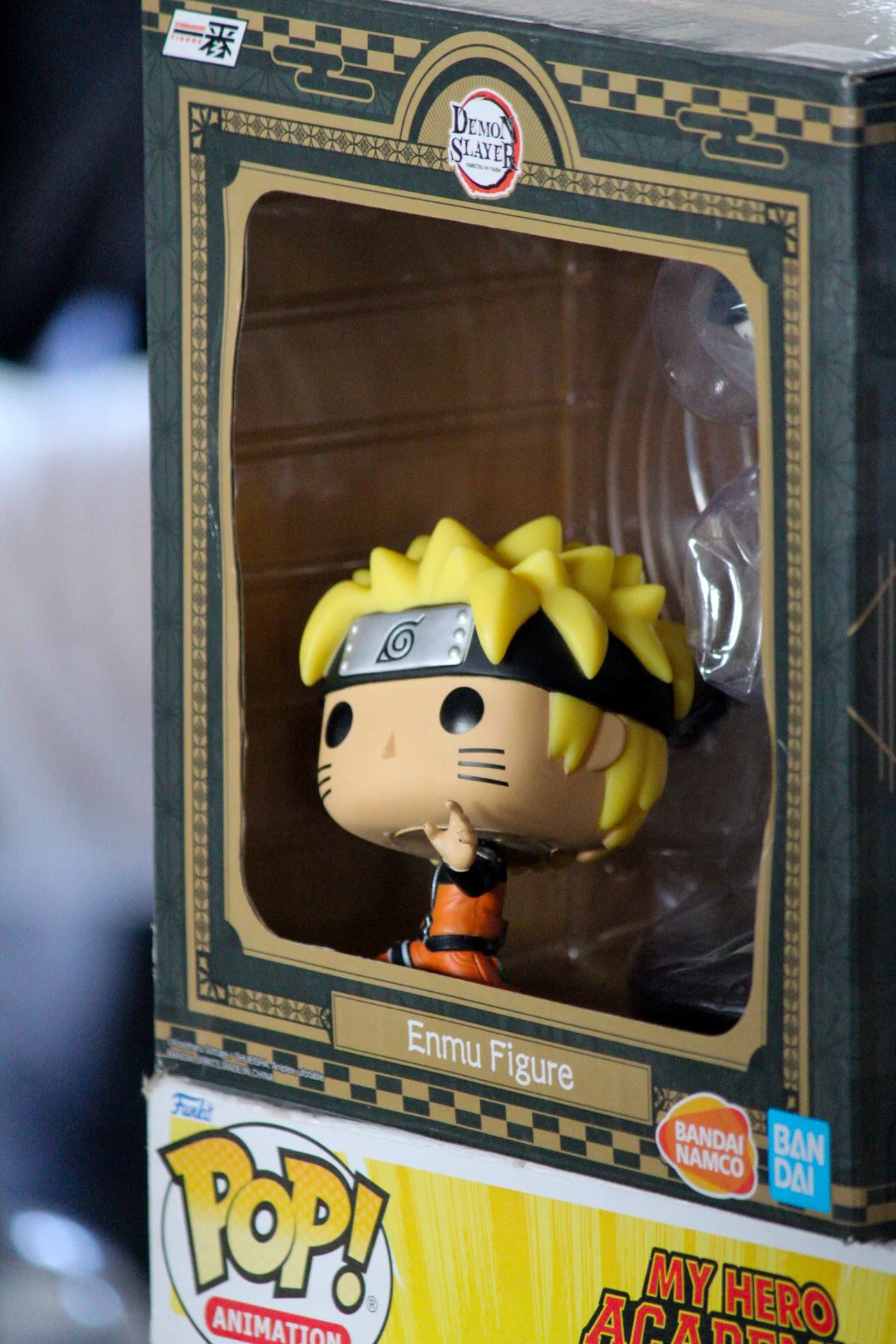 A Funko Pop! Enmu figure from Demon Slayer inside a display box. The figure has yellow hair, a headband with a spiral symbol, and is wearing orange and black clothing. The box has the Demon Slayer logo at the top and is labeled 'Enmu Figure'.
