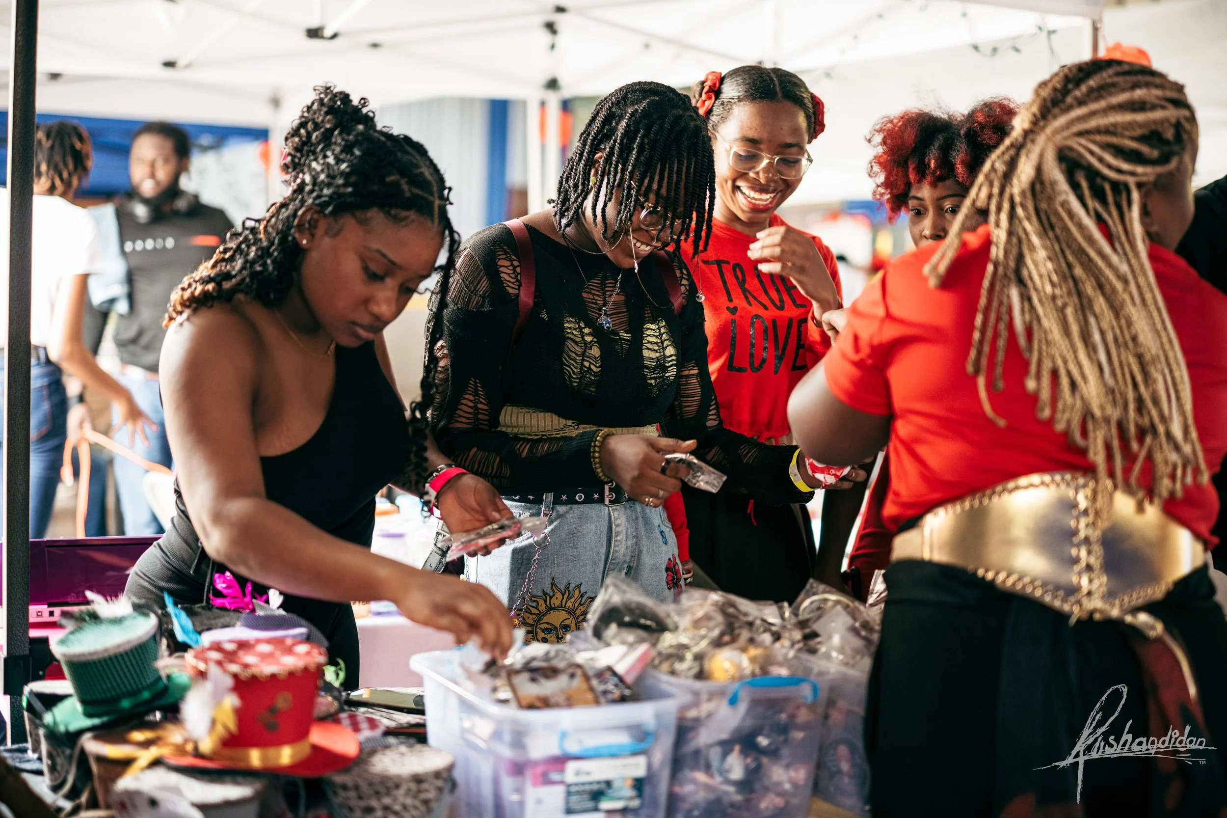 Group of women shopping at an outdoor market stall, looking at various items with smiles.