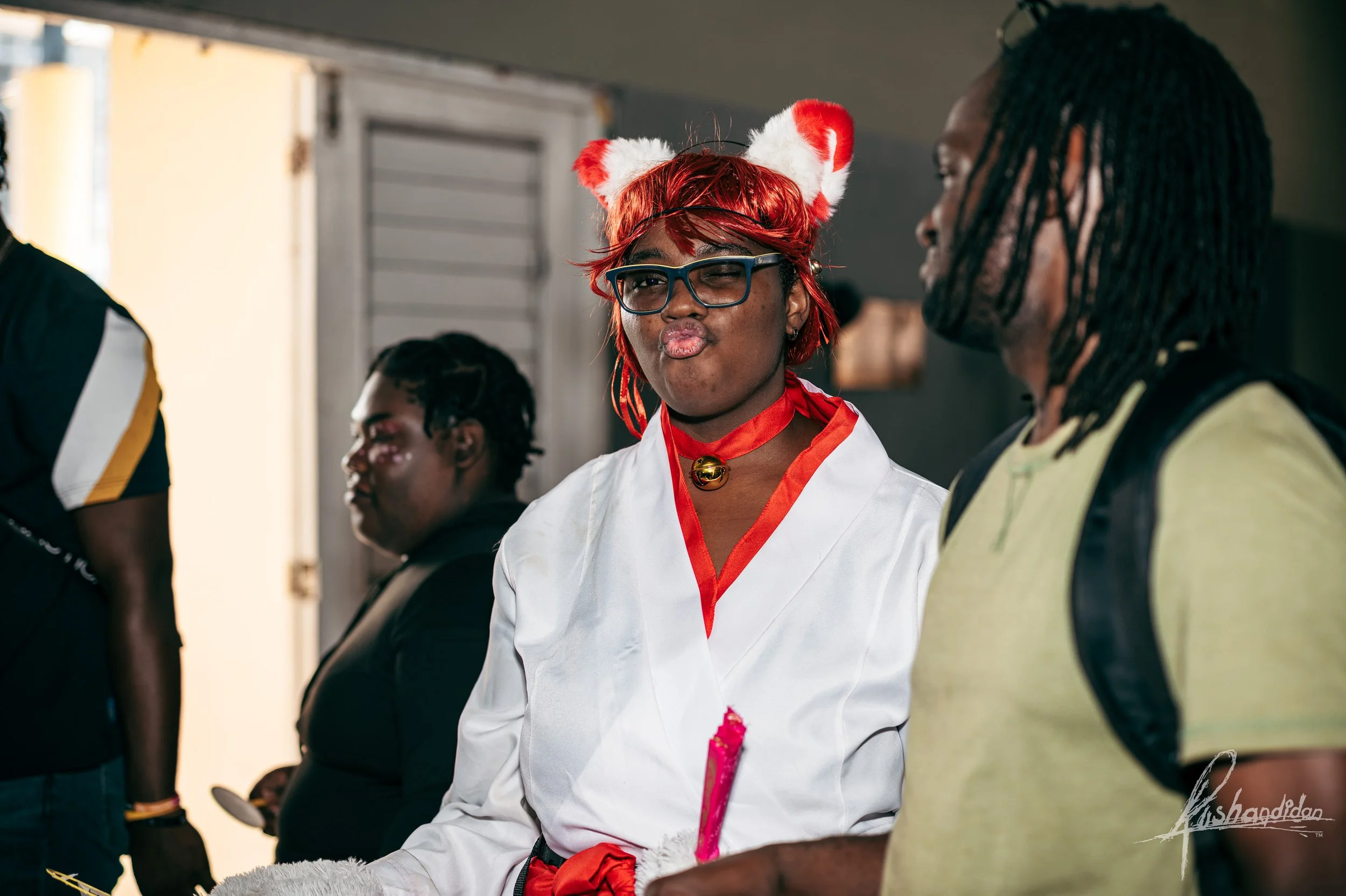 Woman wearing red and white Christmas themed costume with cat ears hat, black glasses, and making a duck face while sitting in an indoor setting with other people around.