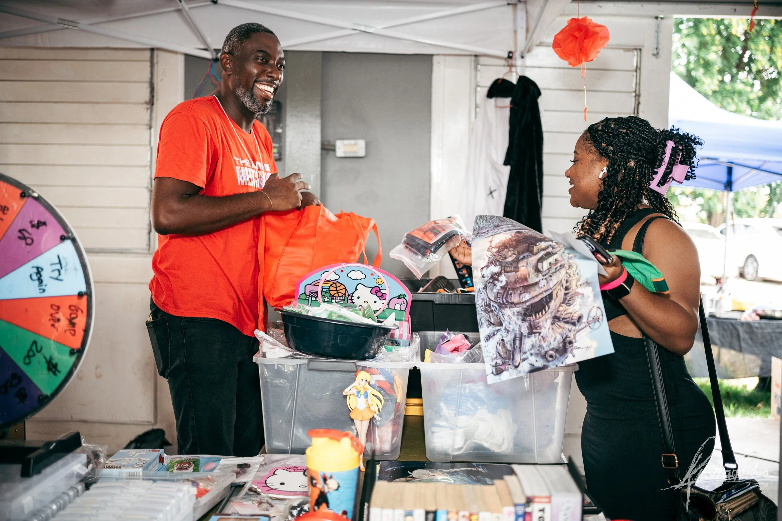 A man and a woman smiling and talking at a booth at an outdoor market, with various toys and merchandise on the table and a spinning wheel in the foreground.